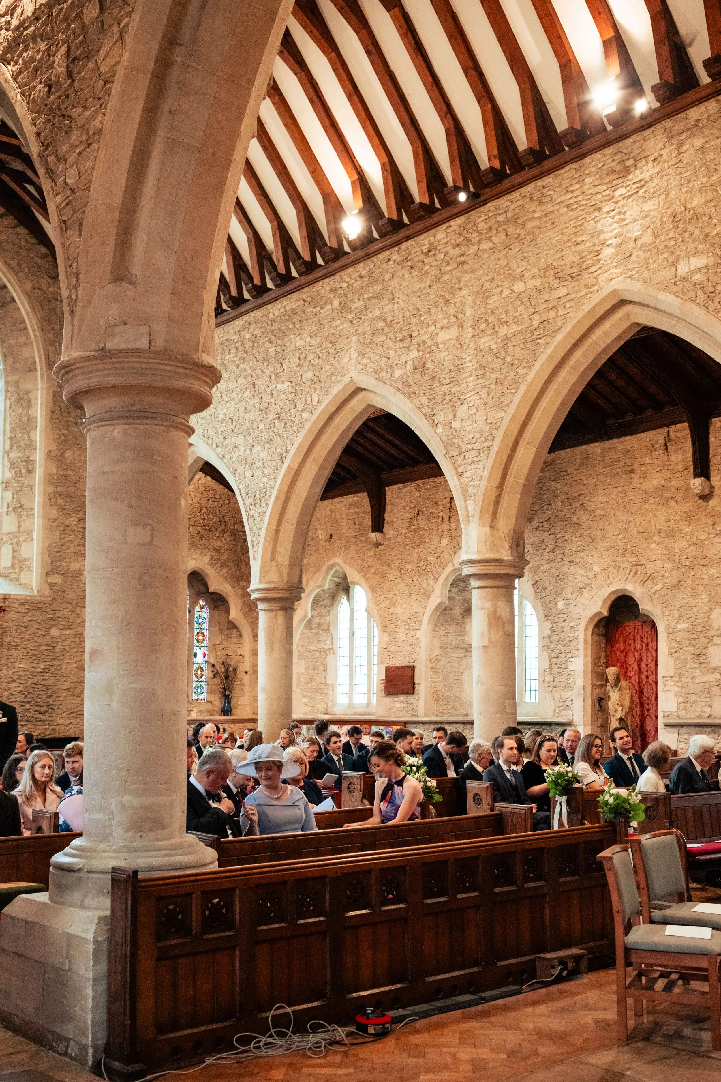 People seated in wooden pews inside a church, attending a wedding, with stone walls, stained glass windows, and Gothic arches.