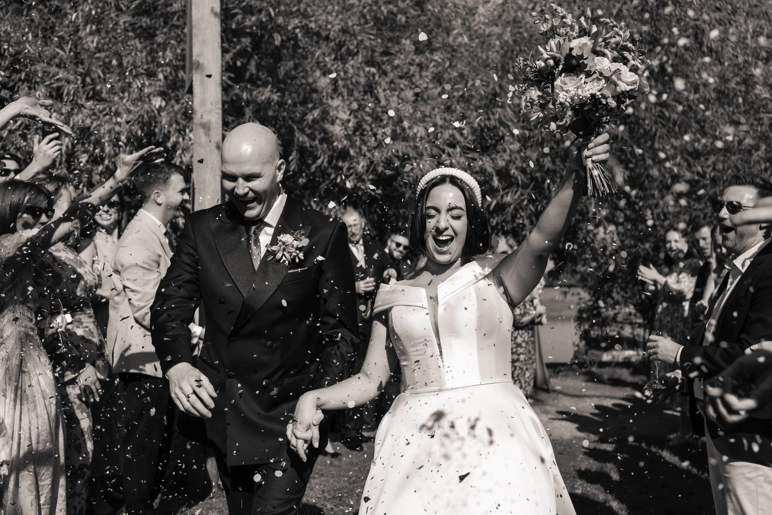 Black and white photo of a newlywed couple celebrating with guests outdoors, with the bride holding a bouquet and everyone throwing confetti.