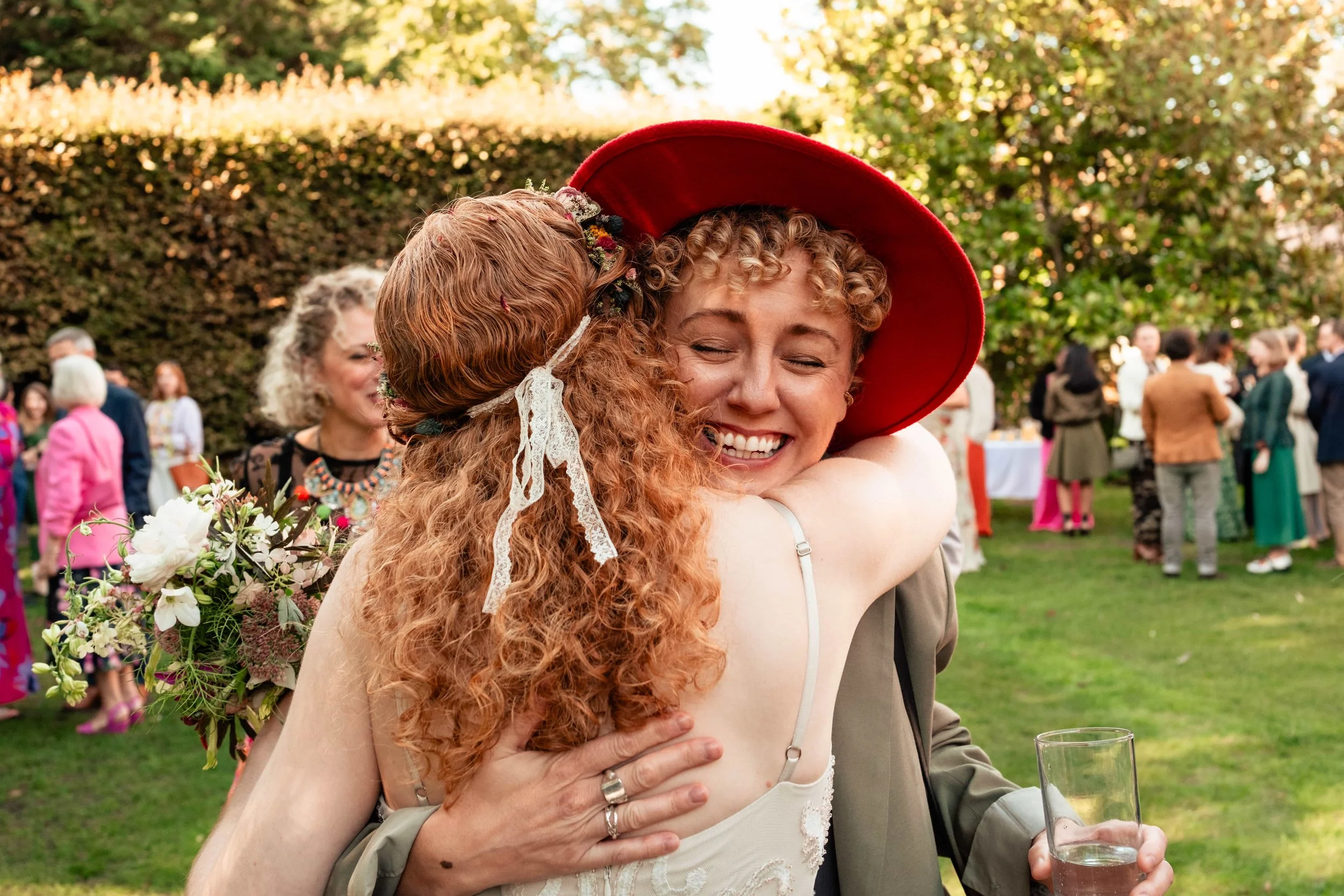 Two women hugging at a social gathering outdoors, one with curly red hair wearing a dress and a floral headband, the other with short curly blonde hair wearing a red hat and a beige blazer, holding a glass of drink, with people mingling in the backgr