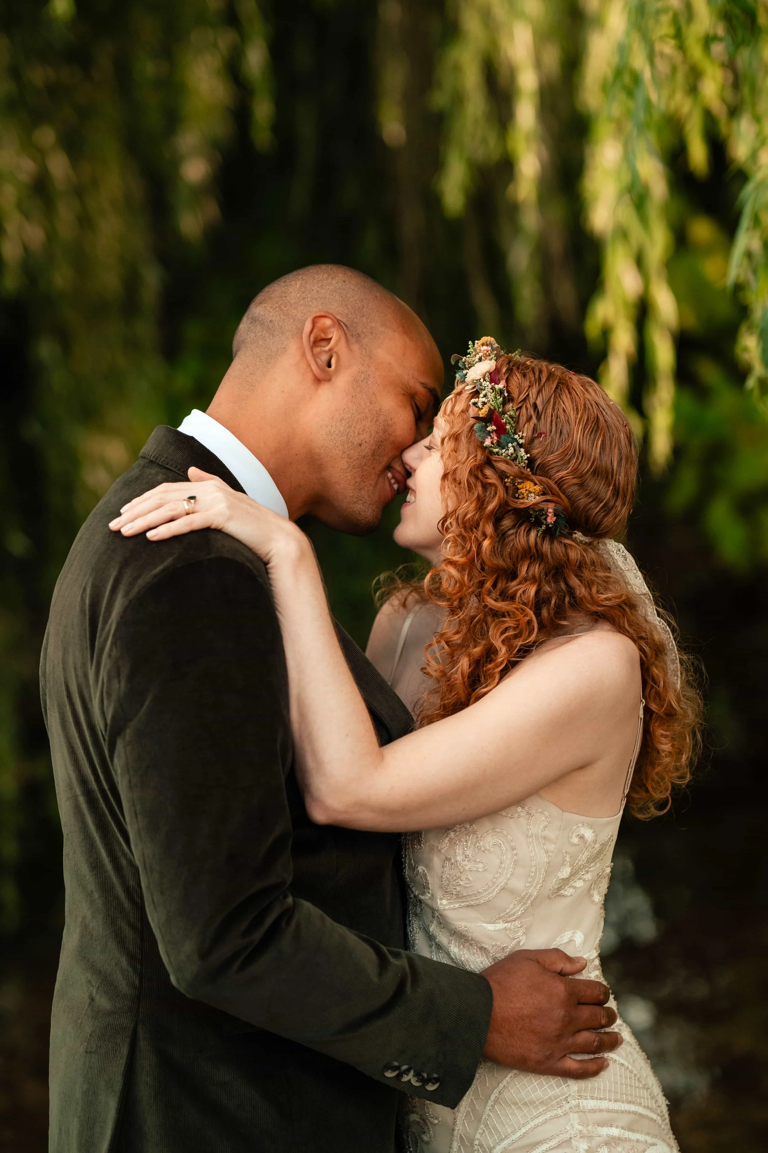 A couple is embracing and touching foreheads in a romantic moment outdoors, with lush greenery in the background. The woman has curly red hair with a floral headband, wearing a lace dress, and the man has short hair, dressed in a black suit.