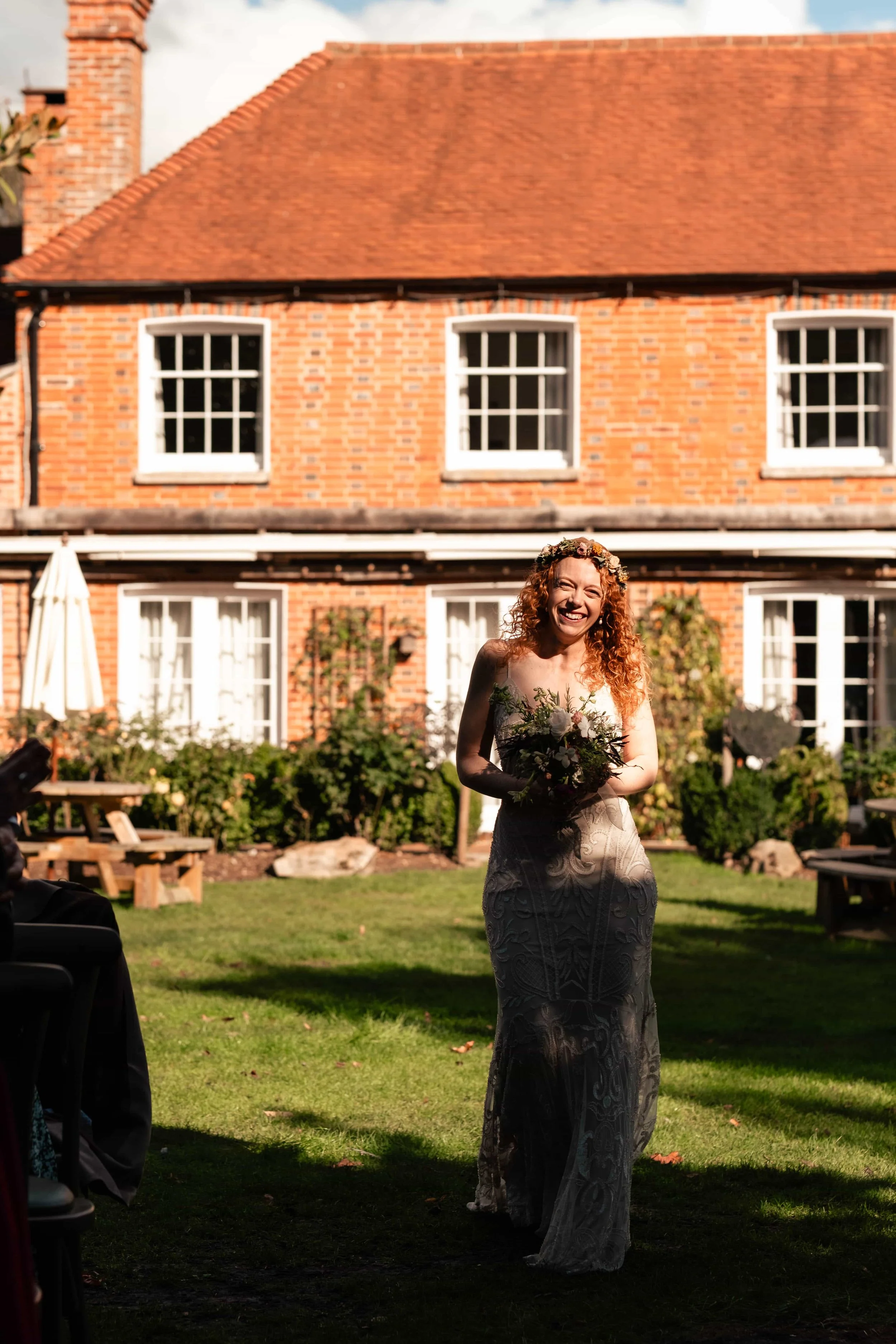 A smiling woman in a lace wedding dress holding a bouquet, standing on a grassy lawn in front of a red brick house with white window frames, during daytime.