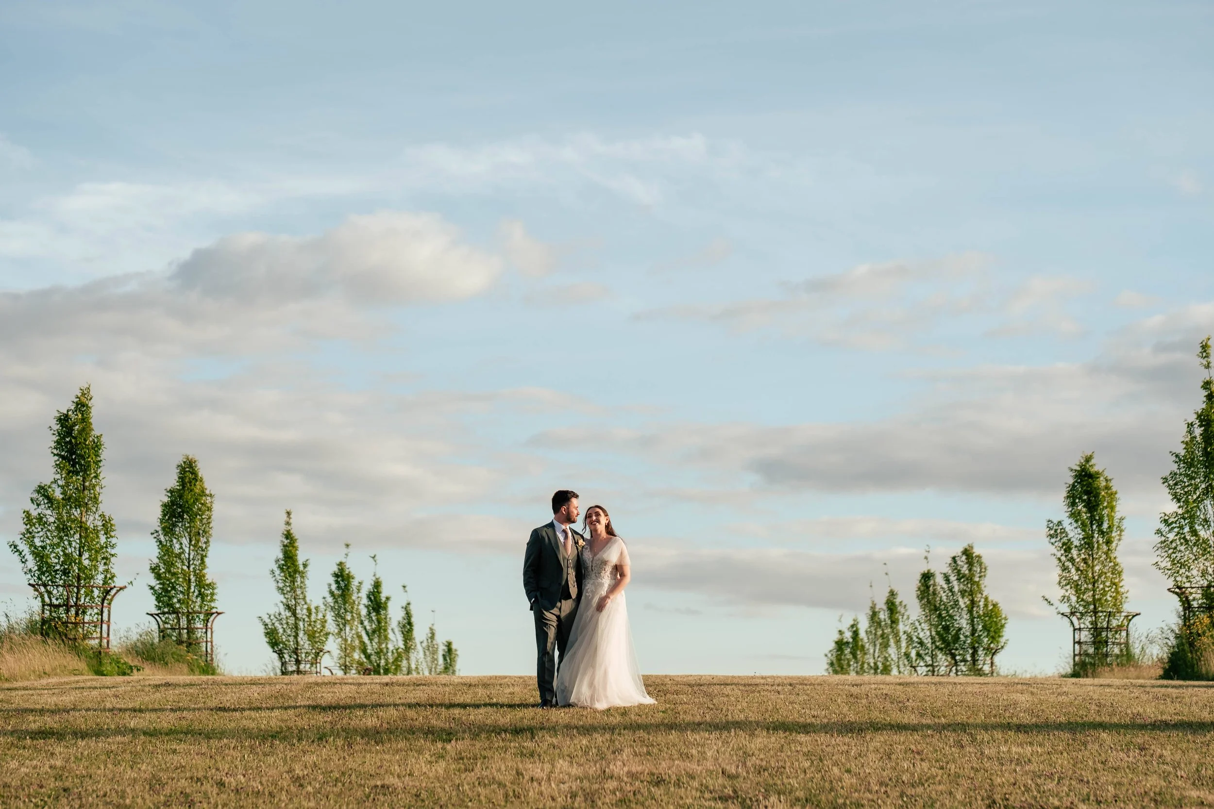 A bride and groom standing on a grassy field with trees and clouds in the background.