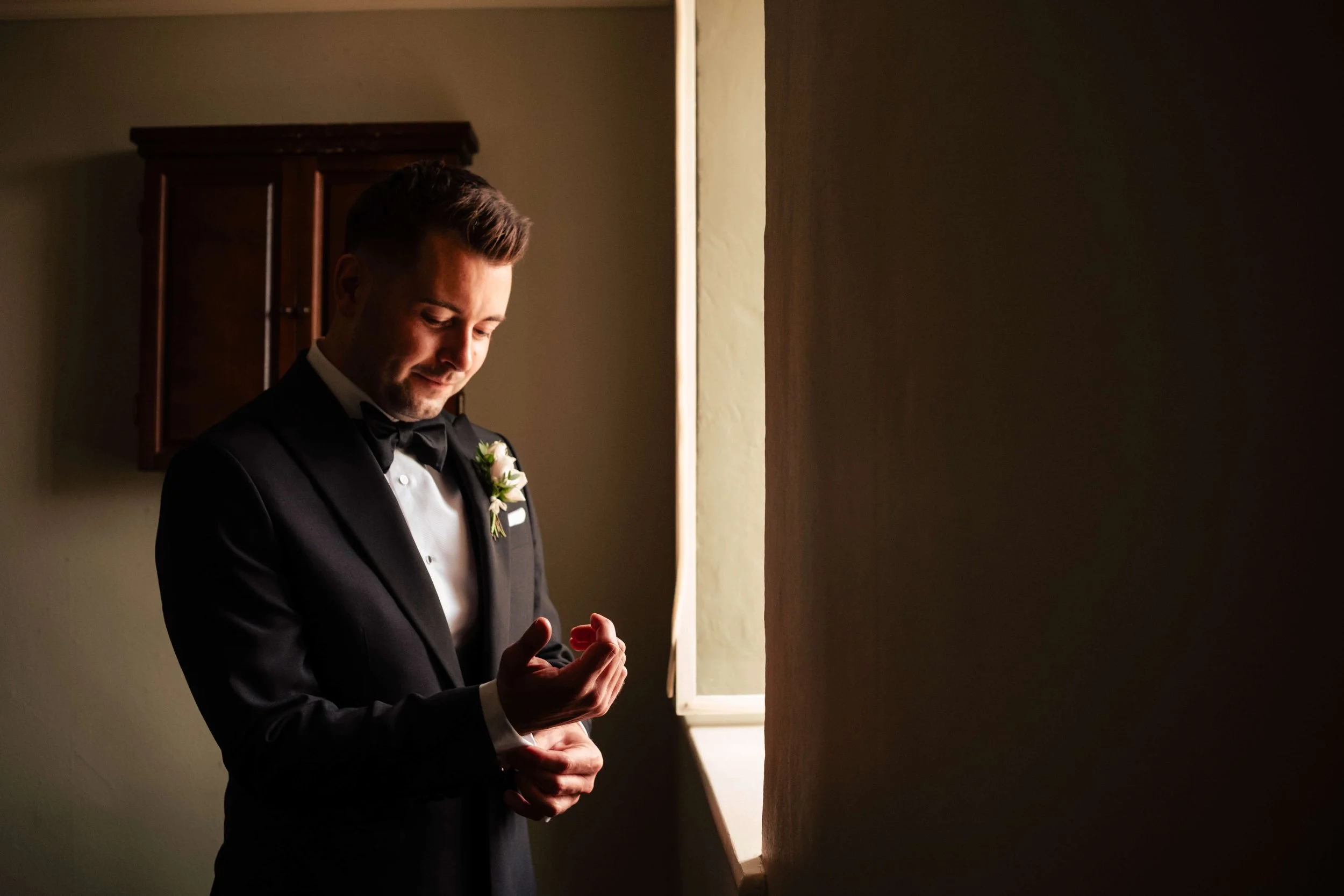 A groom in a tuxedo and bow tie standing near a window, looking down and smiling softly.