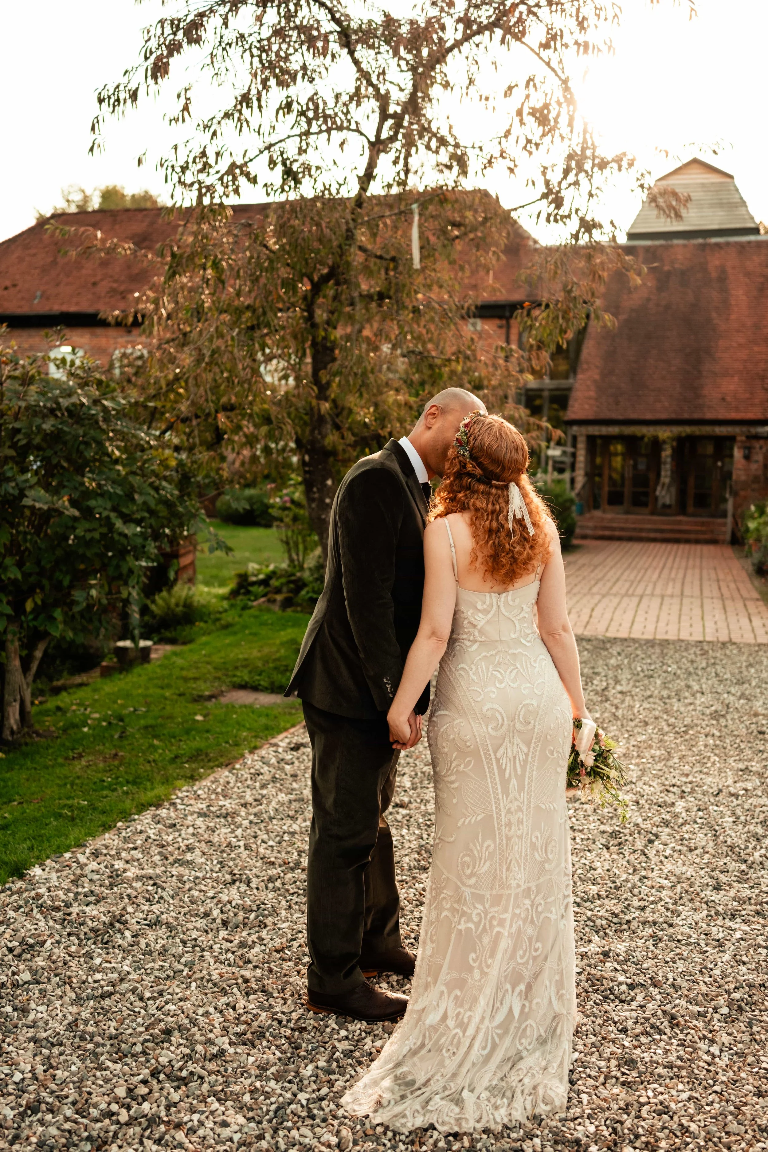 A newlywed couple stands on a gravel path outside a brick house, sharing a kiss at sunset. The bride has red hair, a floral crown, and a white lace wedding dress, and the groom is dressed in a black suit.