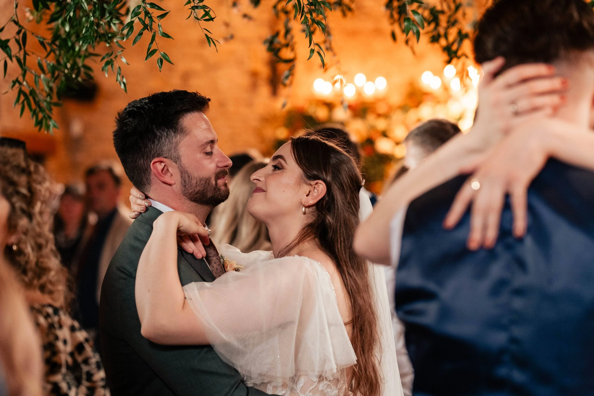 A couple dancing closely at their wedding reception, with guests in the background and warm lighting.