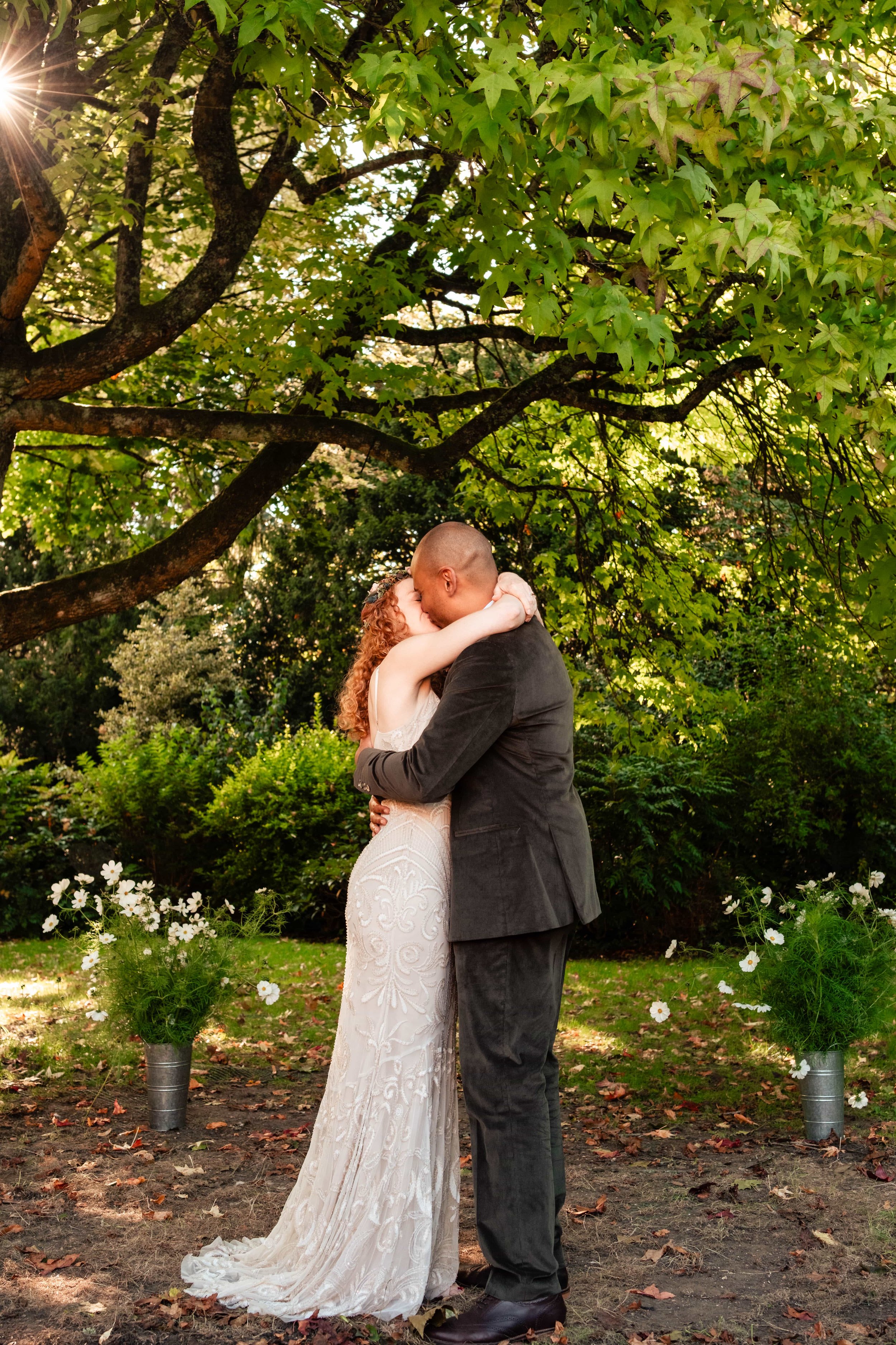 A couple shares a kiss outdoors under a large green-leafed tree during a wedding or romantic photoshoot, with potted white flowers on either side.