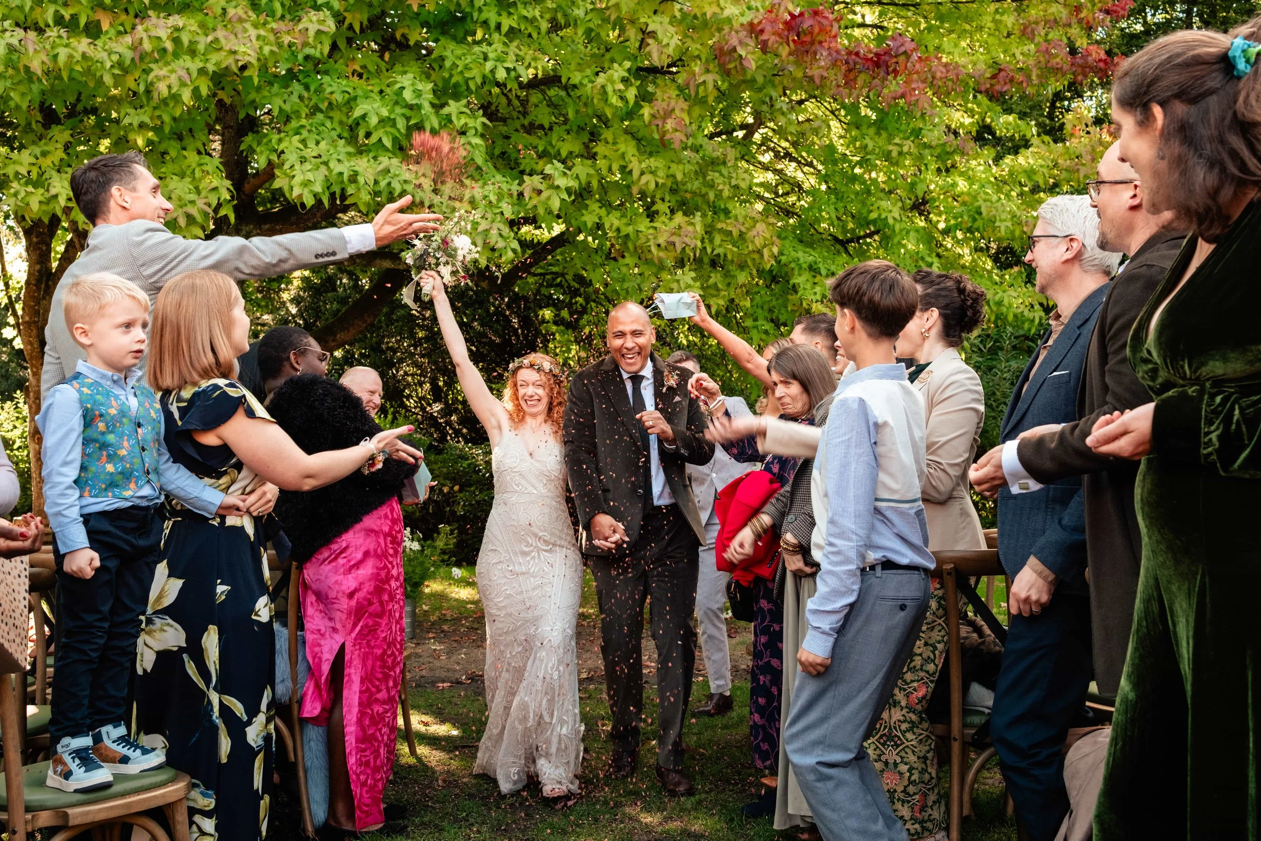 A wedding celebration outdoors with a bride and groom walking hand in hand through a crowd of friends and family. The bride is smiling, wearing a white wedding dress, and the groom in a dark suit. Guests cheer and throw confetti, some reaching out to