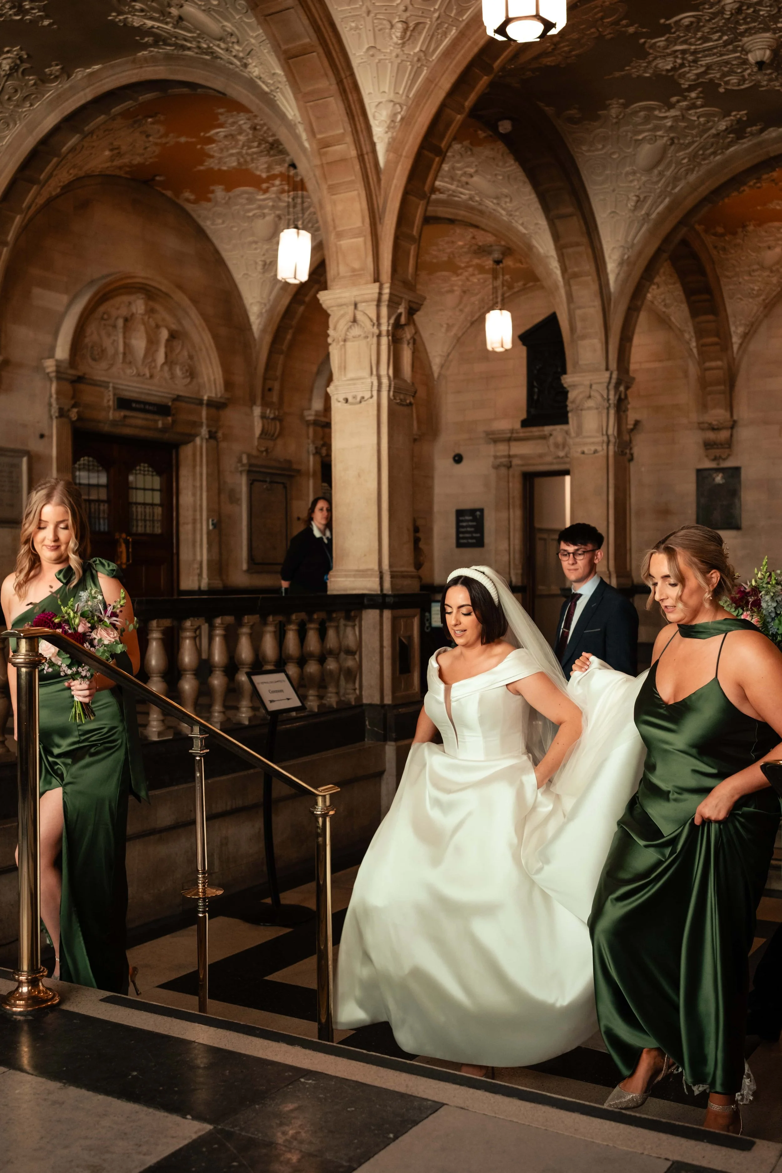 A bride in a white wedding gown walking down a staircase with her bridesmaids in green dresses during a wedding ceremony in the historic walls of Oxford Town Hall.