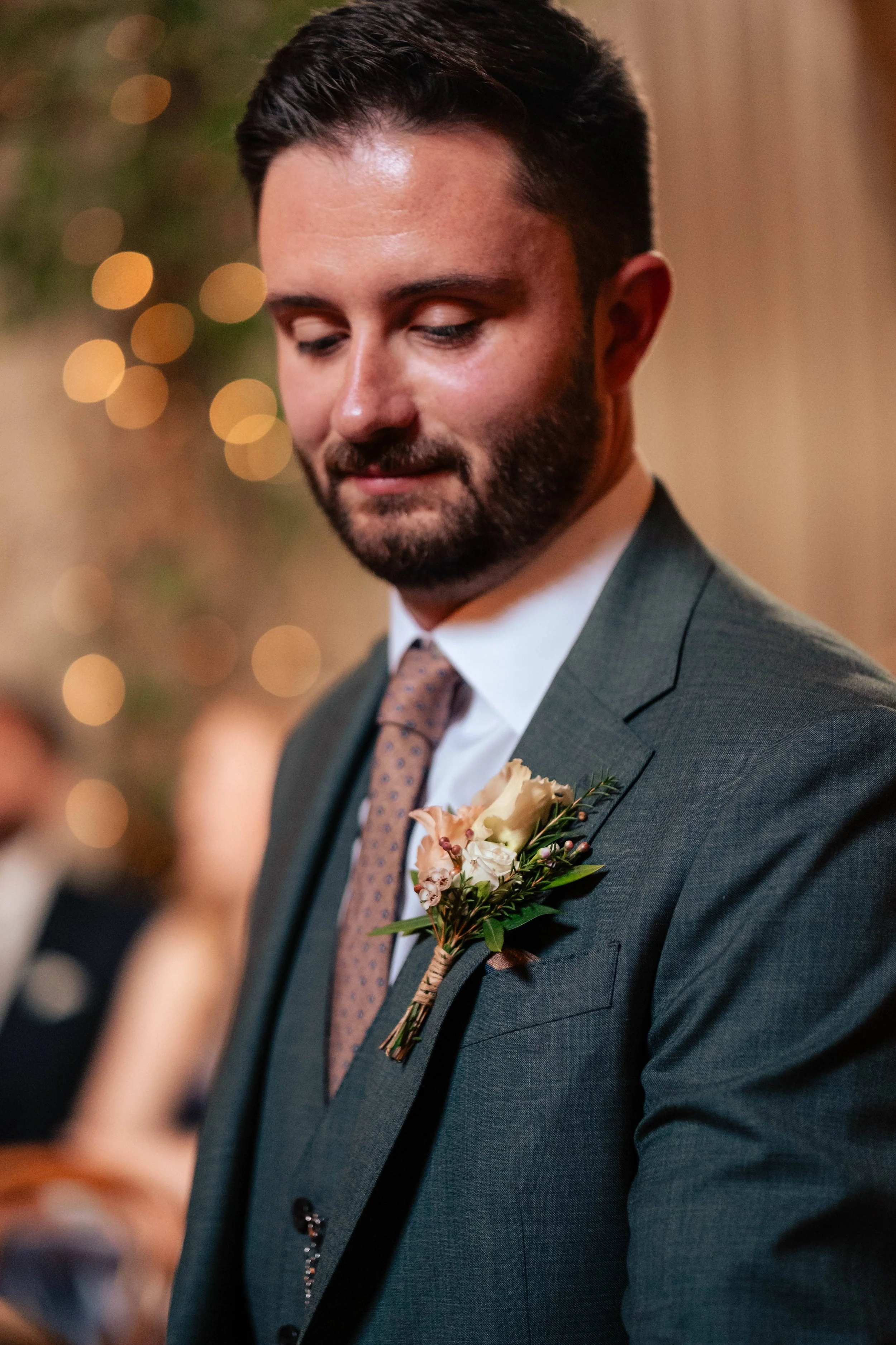 A man in a gray suit with a tie and a boutonniere on his lapel, looking down during a formal event or wedding.