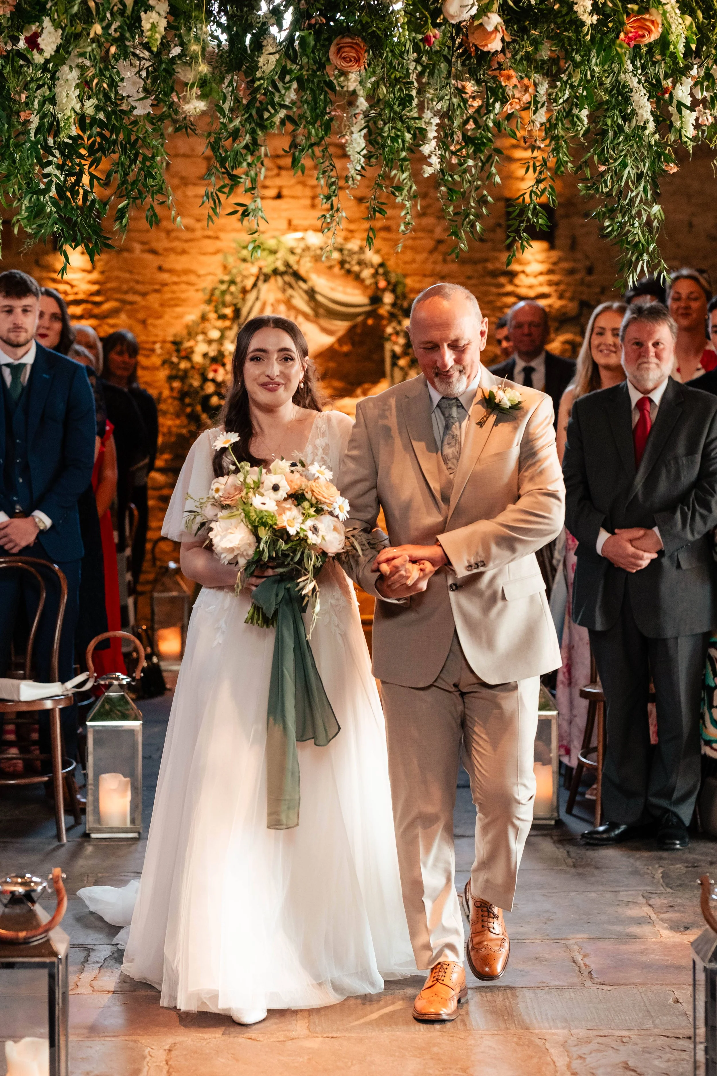 Bride walking down the aisle with her father during her wedding ceremony, surrounded by guests; floral and greenery decorations overhead and candles on lanterns along the aisle.