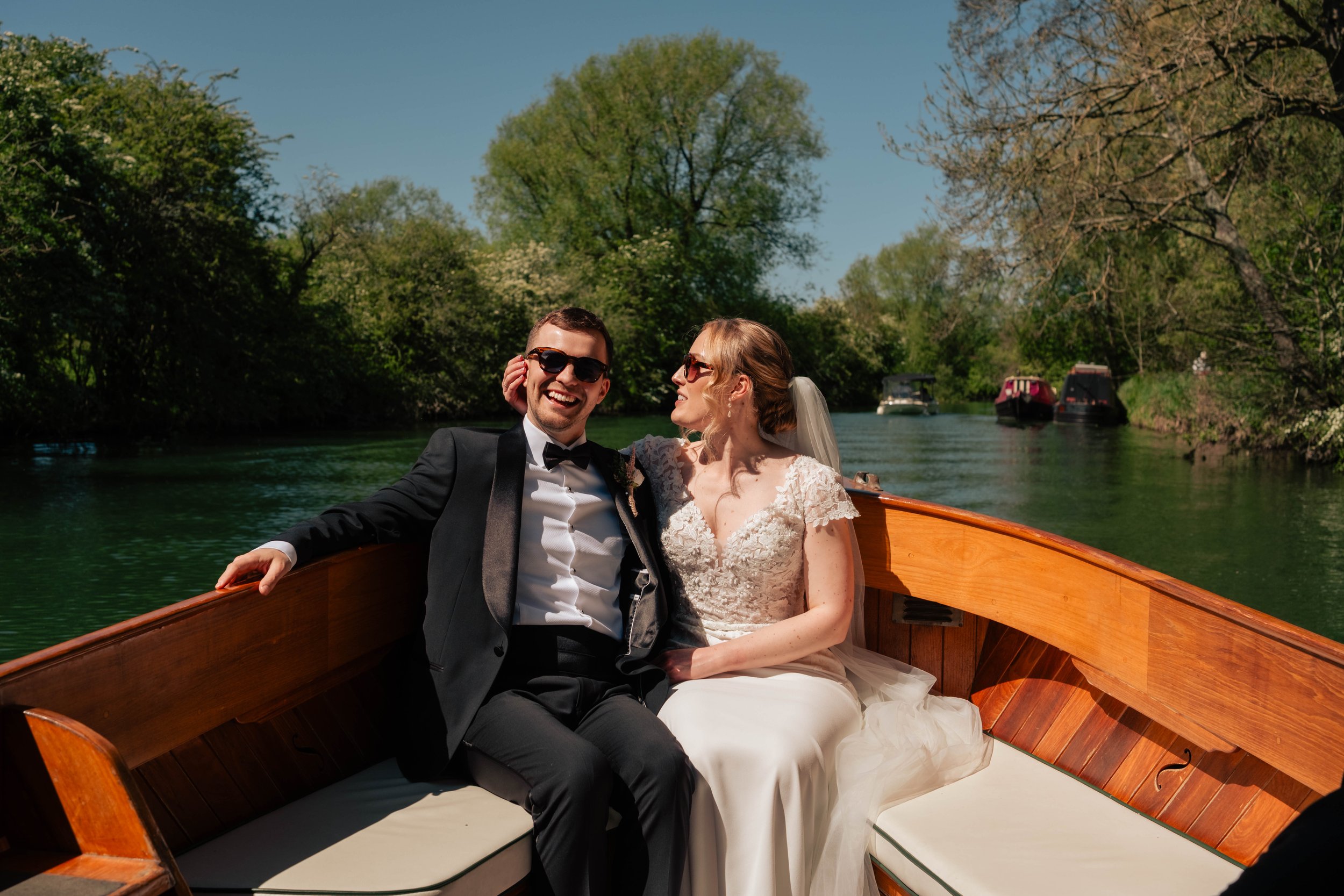 Bride playfully grabbing the groom’s ear as they laugh together at the front of a small river cruiser boat in the sunshine.