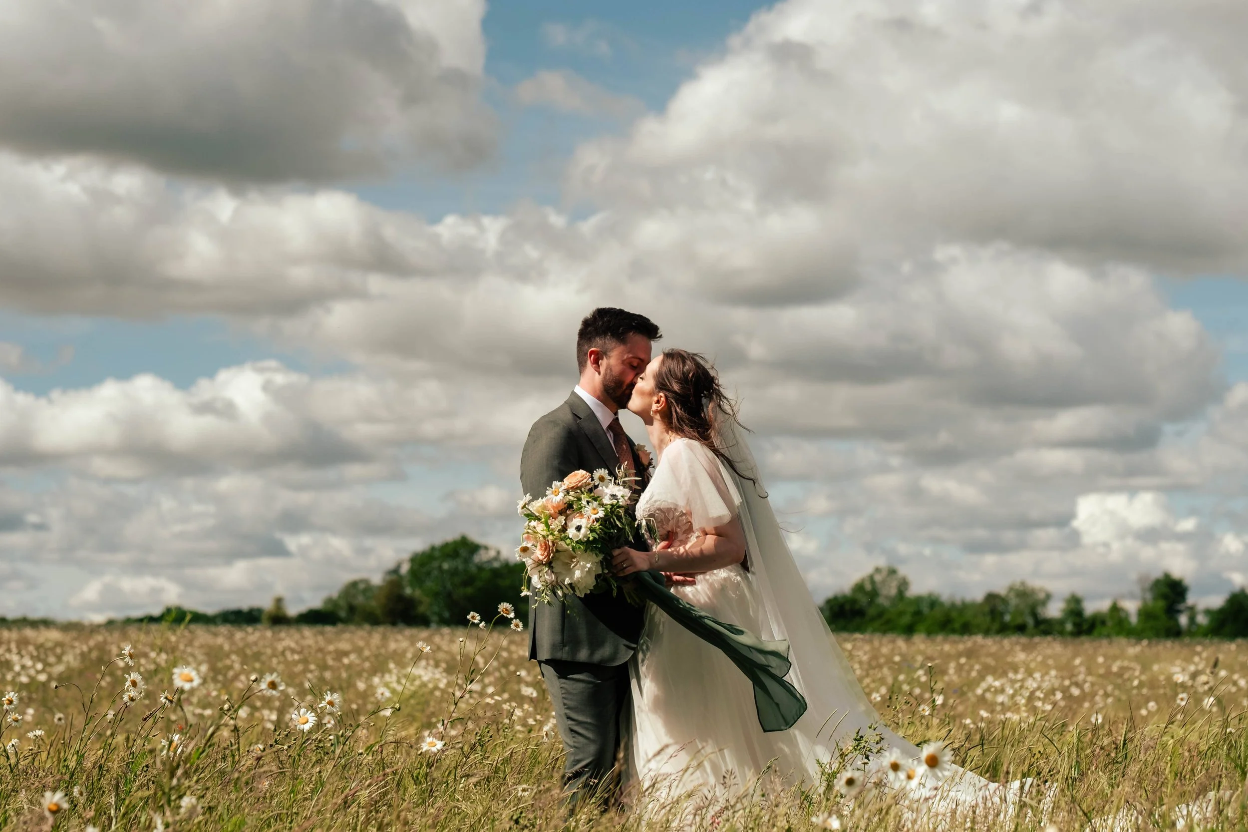 A bride and groom sharing a kiss in a field of wildflowers, with a backdrop of a cloudy sky and trees in the distance.