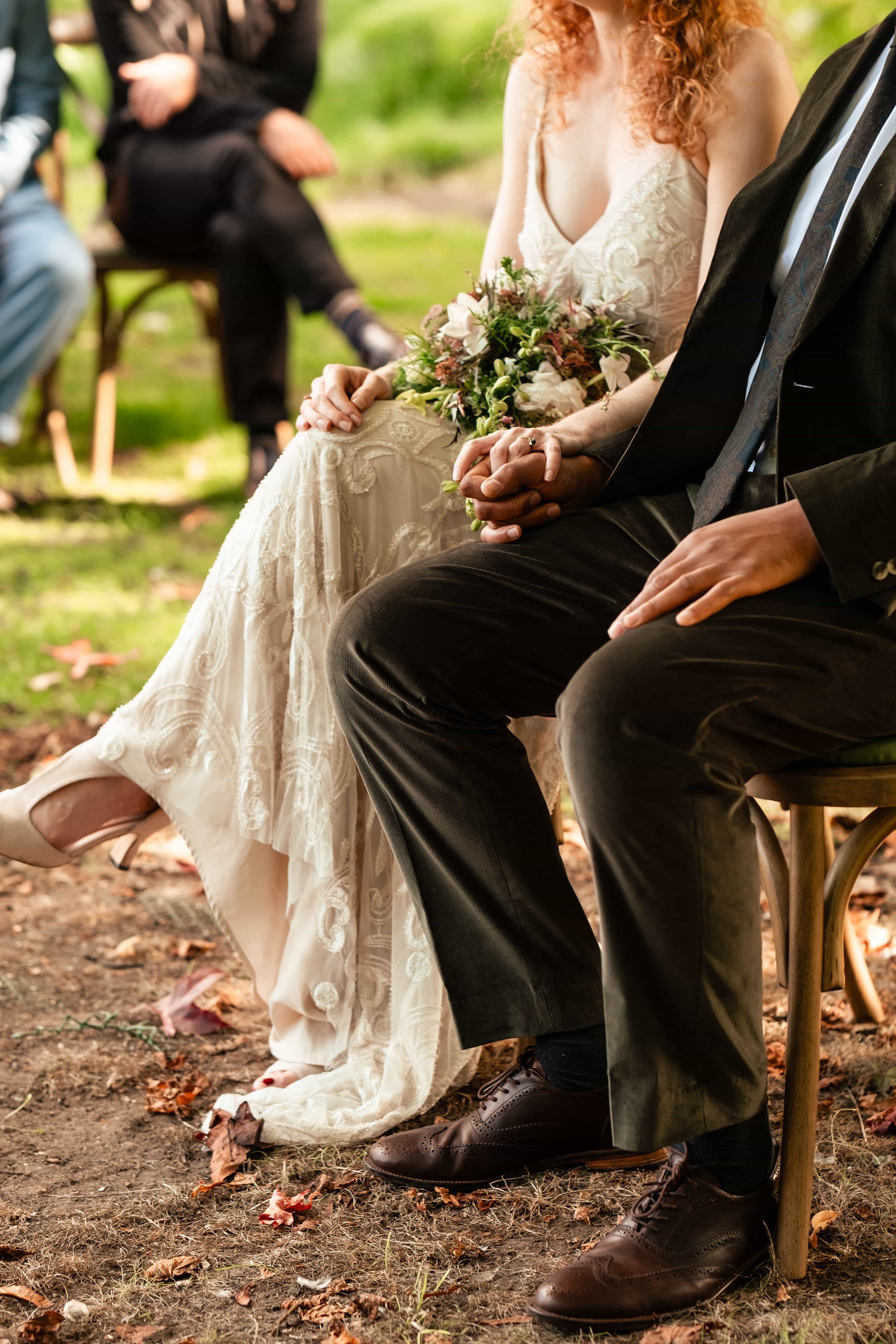 Close-up of a bride and groom sitting hand-in-hand at an outdoor wedding, with guests in the background.