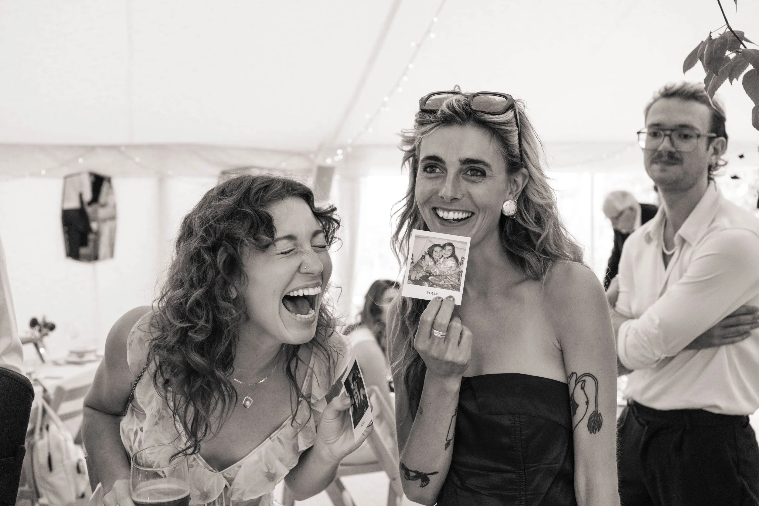Two women laughing and holding a photograph at a social gathering, with a man observing in the background.