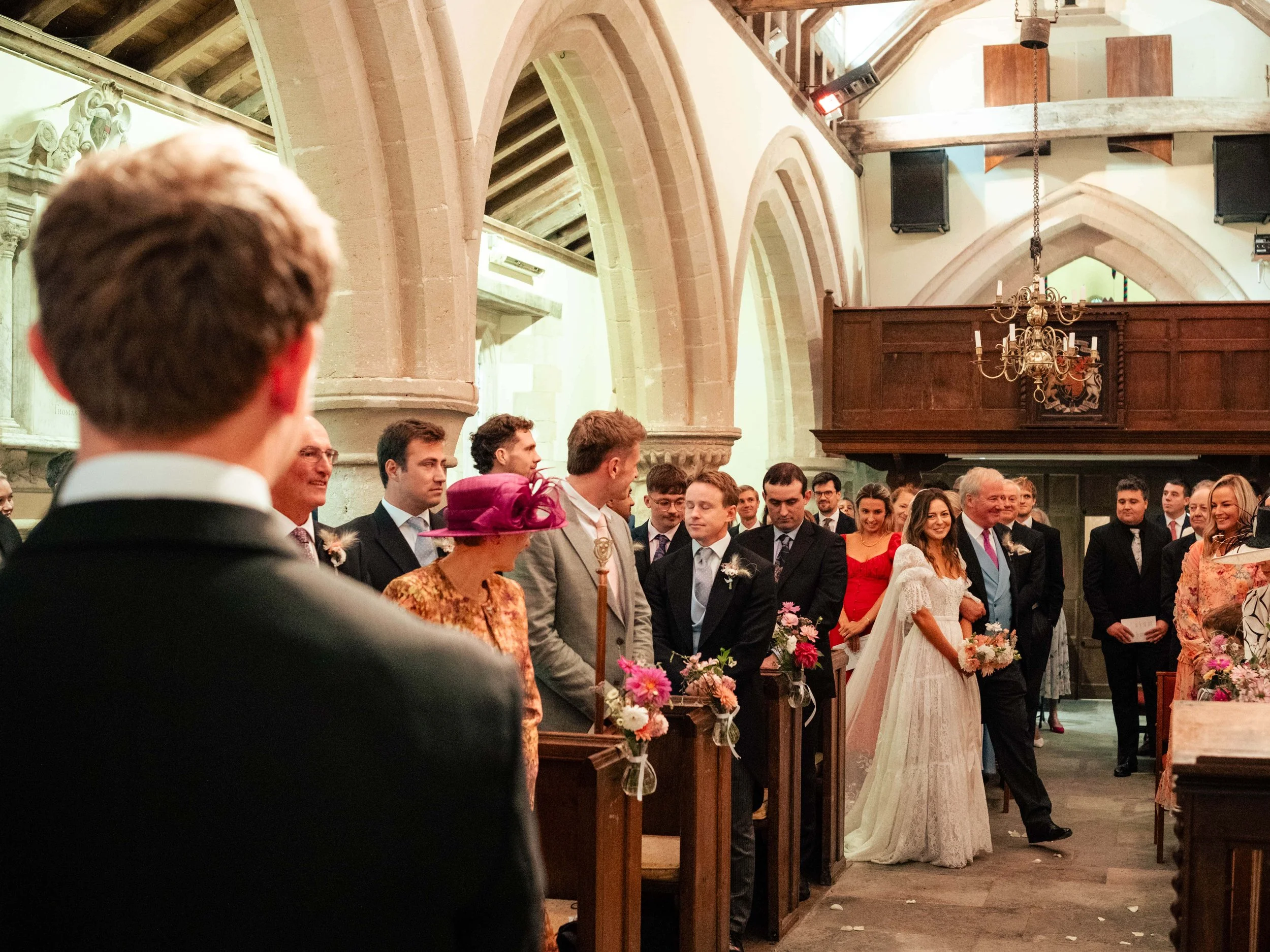 Wedding ceremony inside a church, with the bride in a white gown holding a bouquet, surrounded by guests dressed in formal attire.