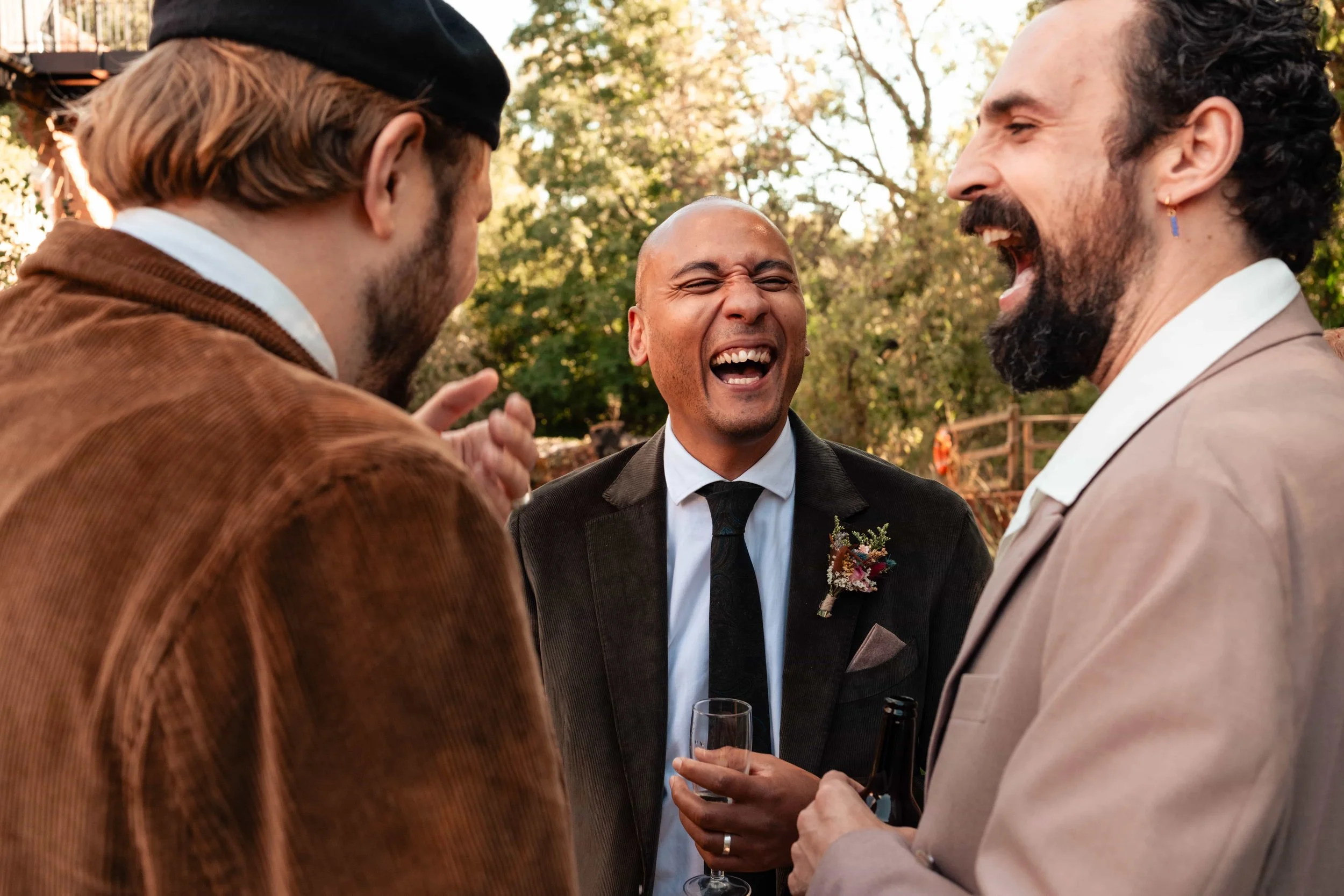 Three men laughing and talking at an outdoor social event, with one holding a glass of champagne and another holding a bottle, during daytime with trees in the background.