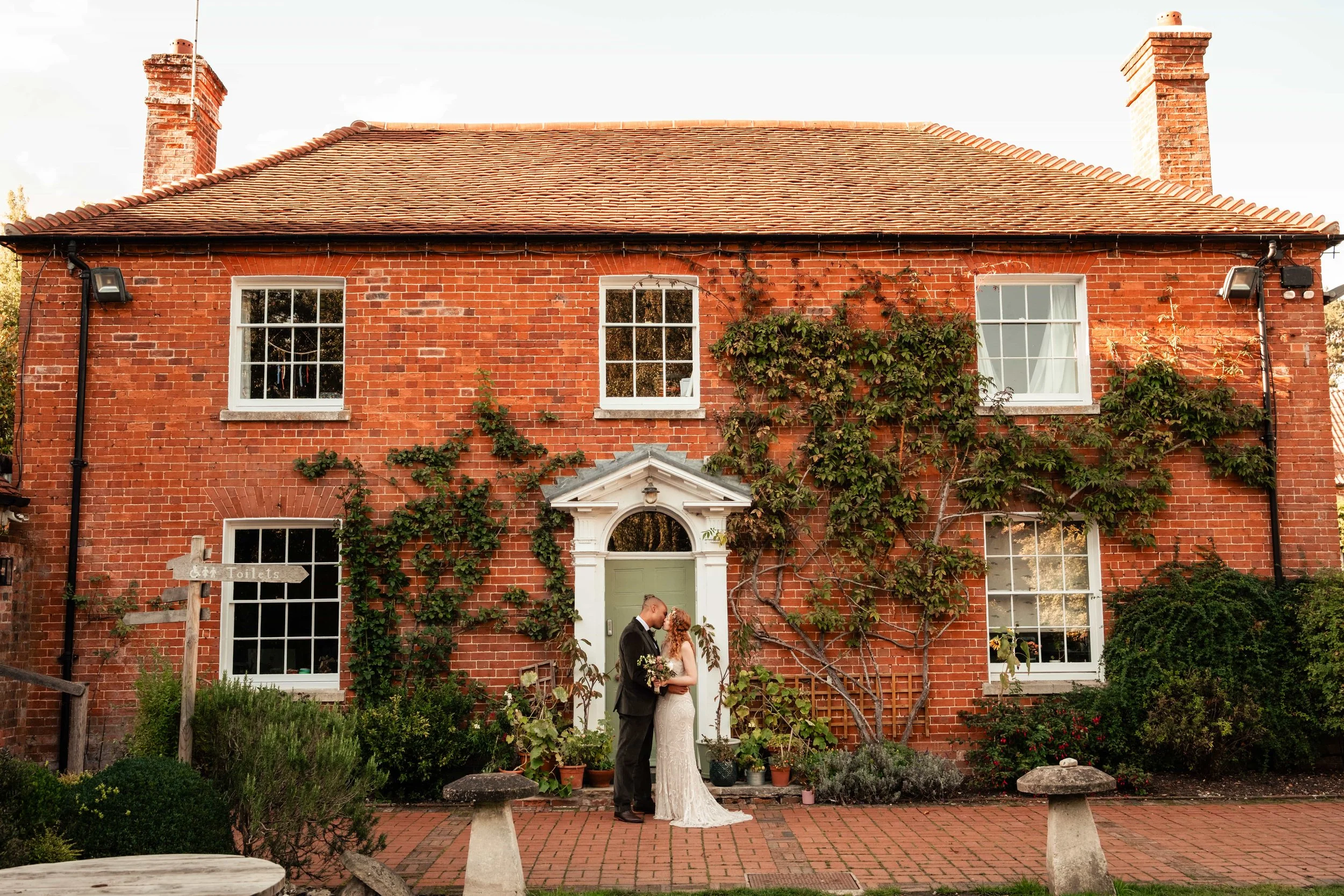 A couple, dressed in wedding attire, standing close together in front of a brick house with greenery and ivy climbing on the walls.