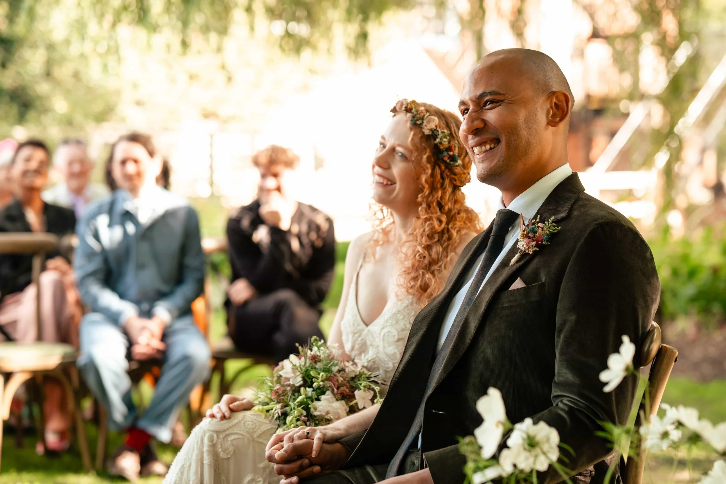 A bride and groom sitting outdoors at their wedding ceremony, smiling and looking at each other, with guests seated behind them.