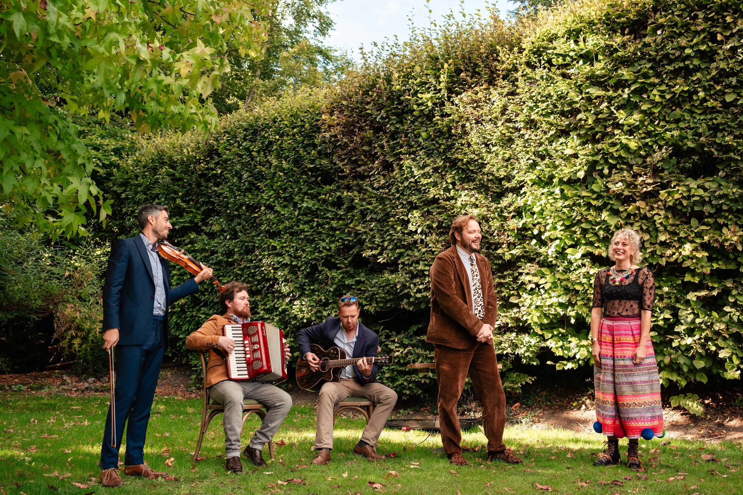 Five people outdoors in front of a large green hedge, some playing instruments and others standing and smiling. One man is playing a violin, another is playing an accordion, and a third is playing a guitar. The two standing individuals are a man and 