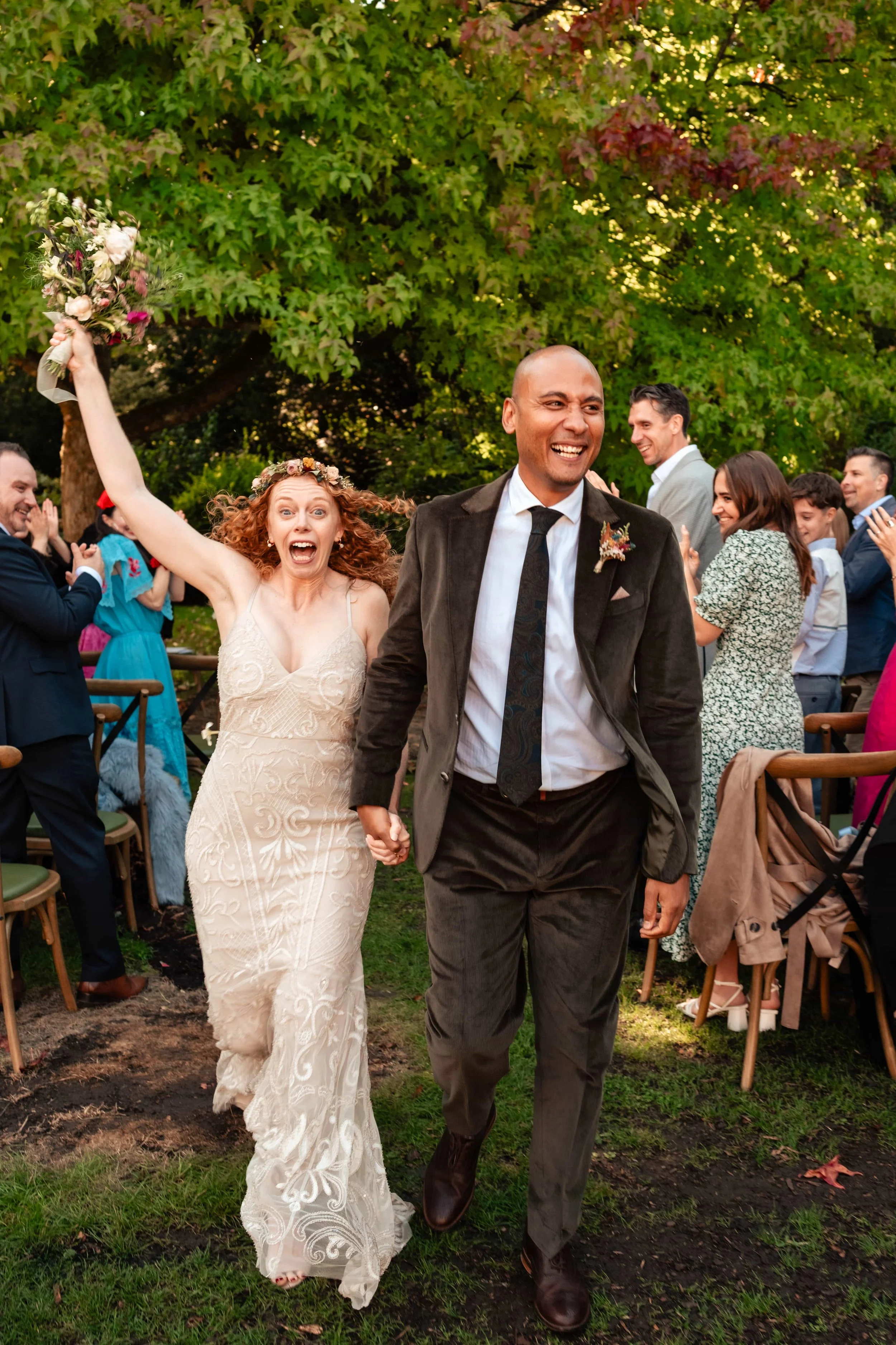 A joyful newlywed couple holding hands and walking down the aisle outdoors at a wedding reception, with guests clapping and smiling in the background.