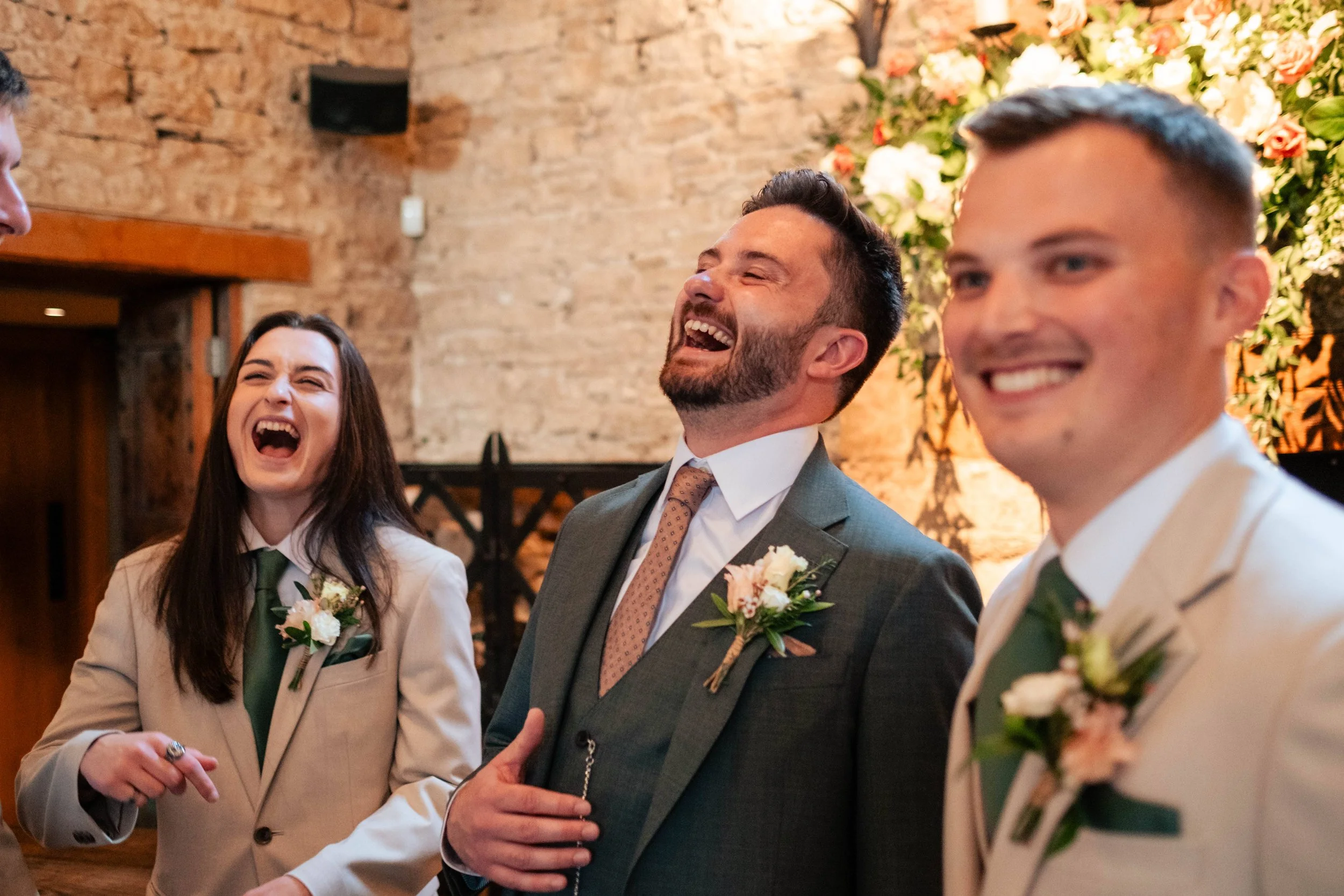 Three people, two men and one woman, dressed in formal suits with boutonnieres, sharing a laugh at a wedding celebration indoors with a brick wall and floral decorations in the background.