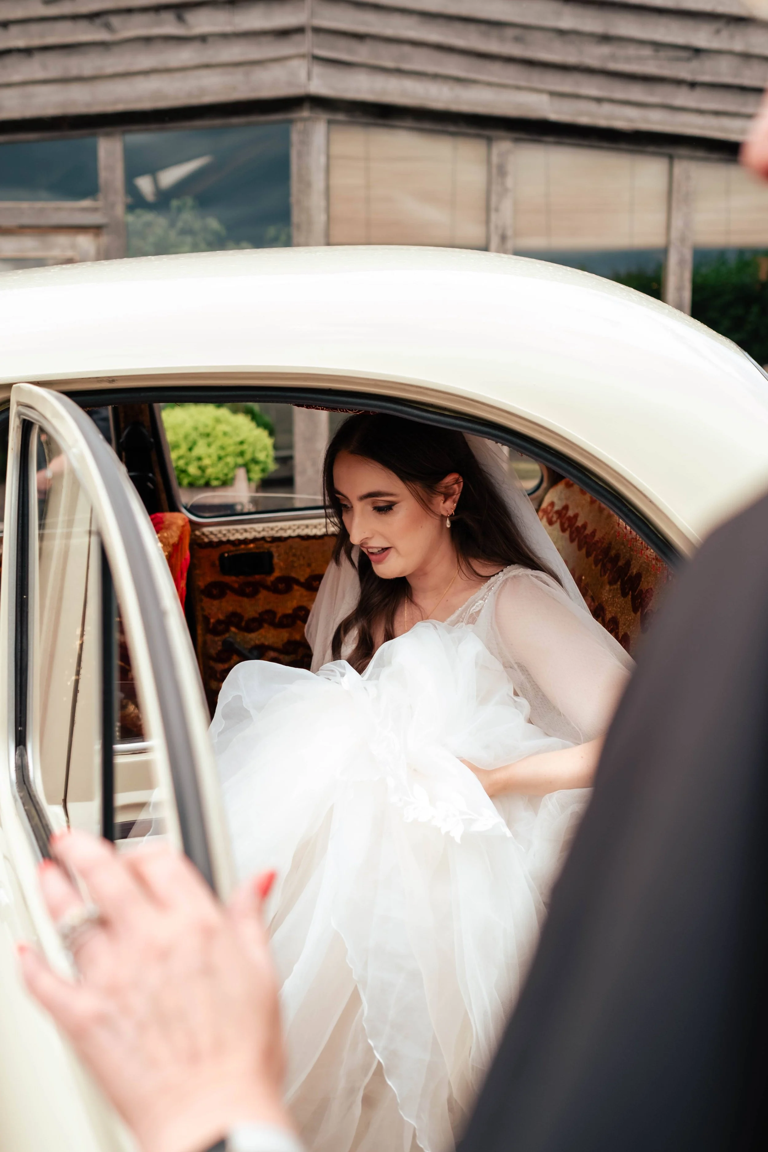 Bride in a white wedding dress sitting in a vintage car, looking down, with a person outside the car touching the door.