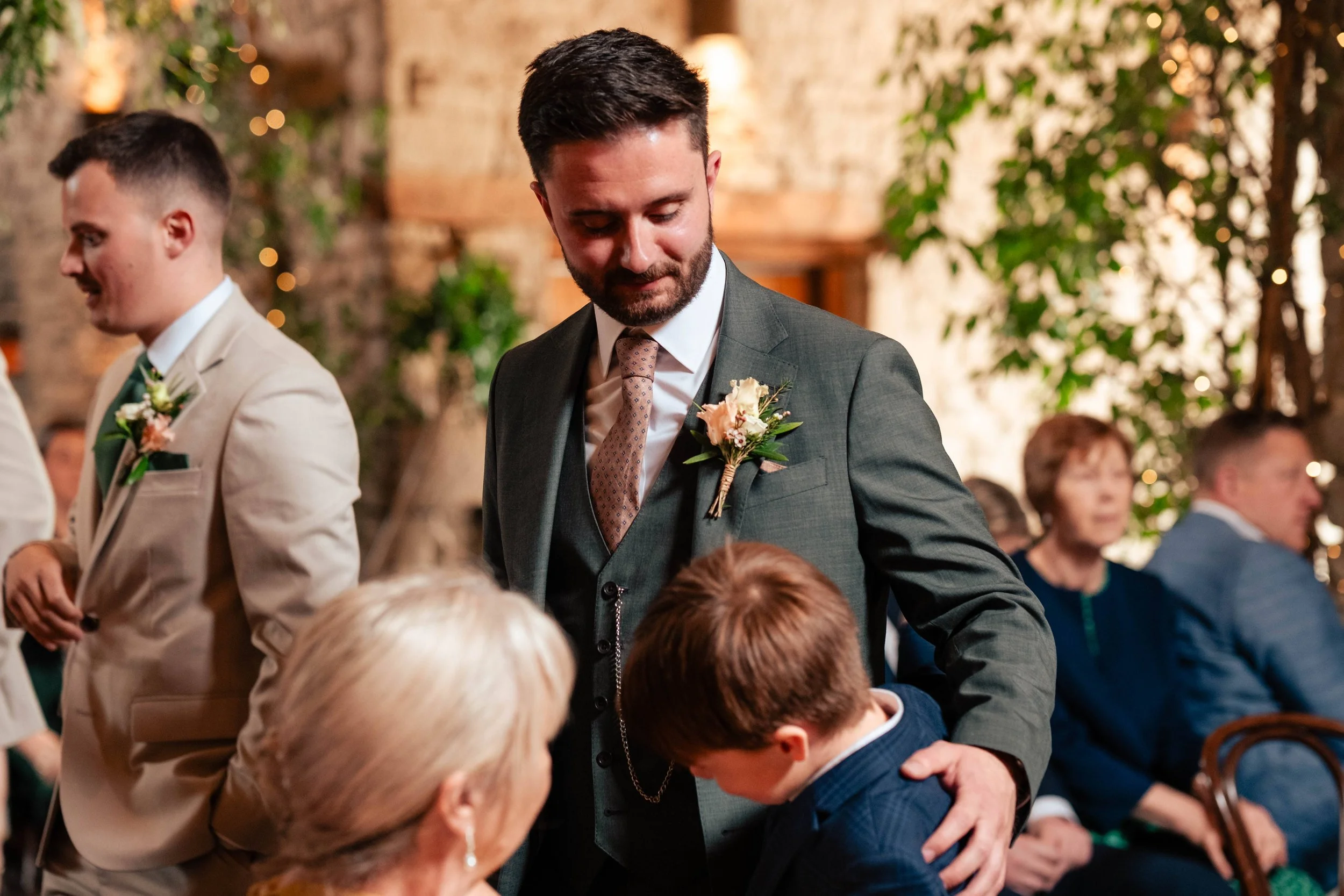 A man in a gray suit with a pink boutonniere gently interacts with a young boy at a wedding reception, while other guests are seated in the background.