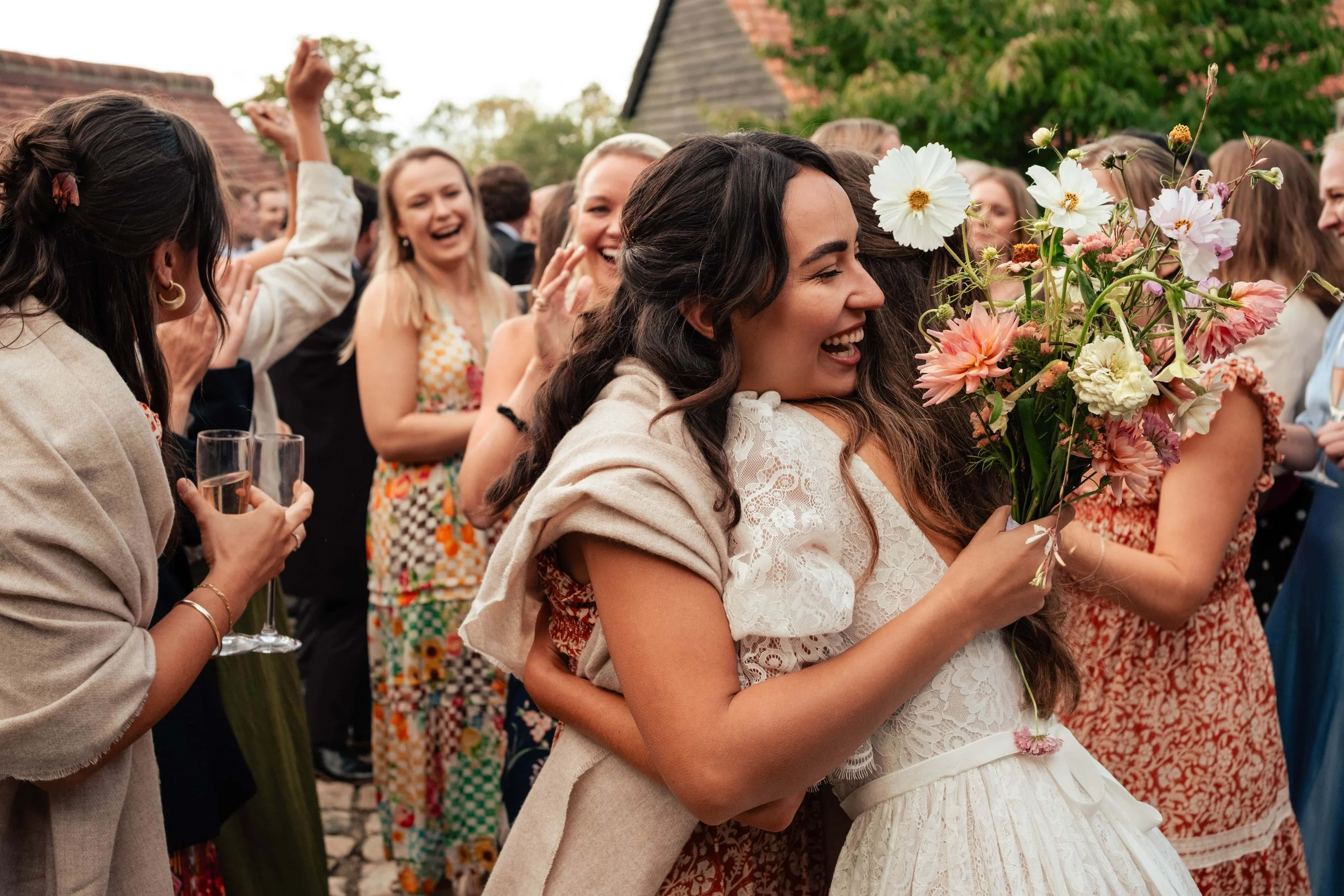 A woman in a white lace dress is holding a colorful bouquet of flowers, smiling, while being embraced from behind by another woman during a celebration with a group of smiling people.