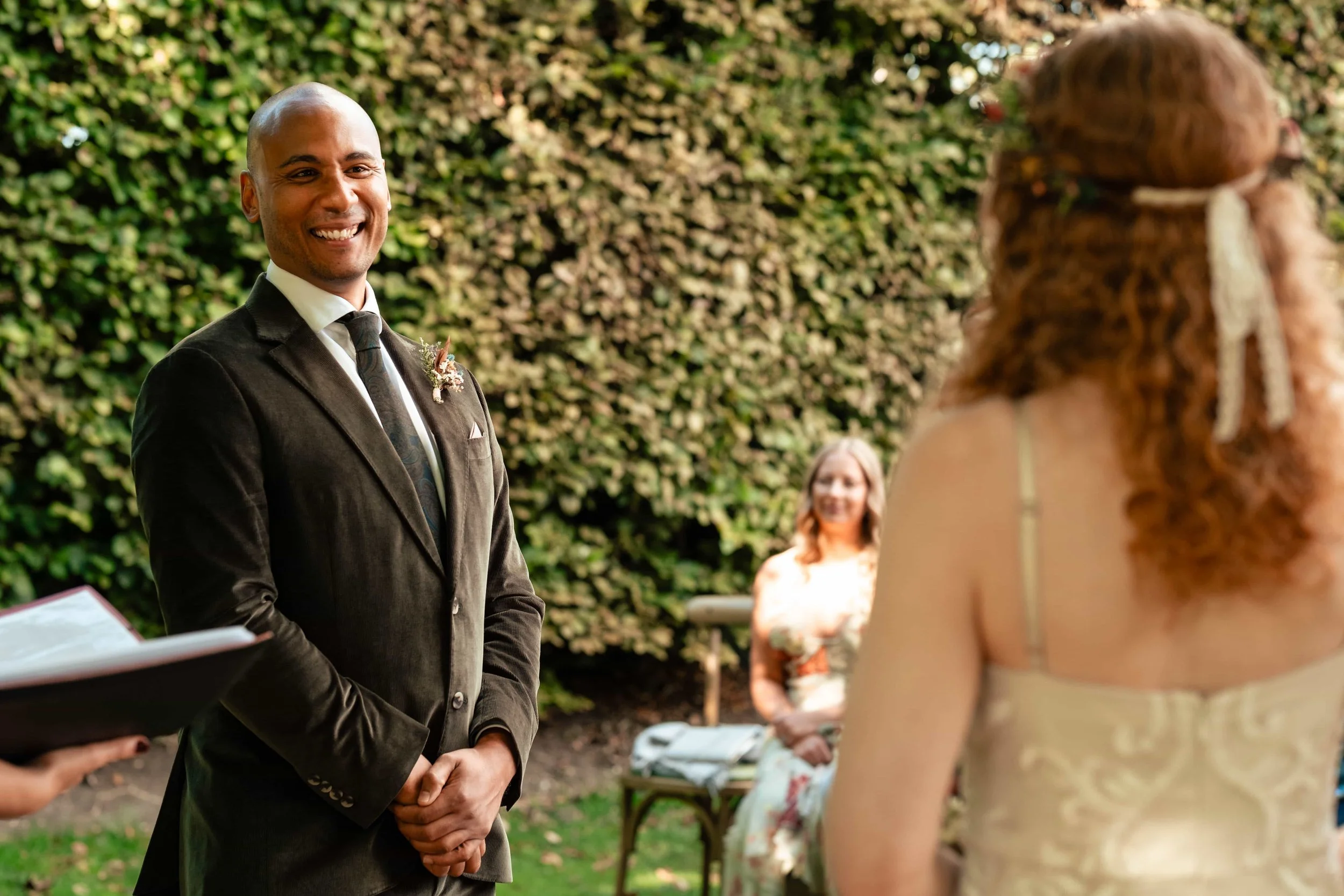 A groom in a dark suit and tie stands smiling at a wedding ceremony outdoors in front of a green leafy background, with a bride in a white dress with lace details and curly hair with a ribbon, and a woman seated in the background.