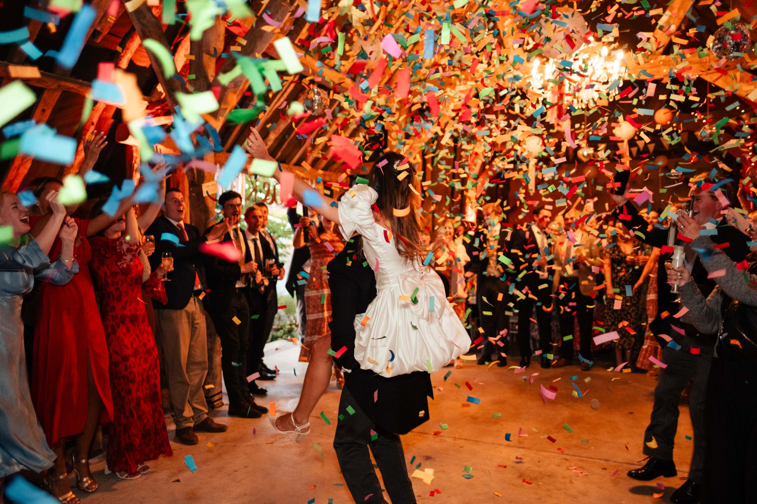 People celebrating at a wedding reception with confetti falling, a bride in a white dress dancing with a woman, surrounded by guests clapping and smiling.