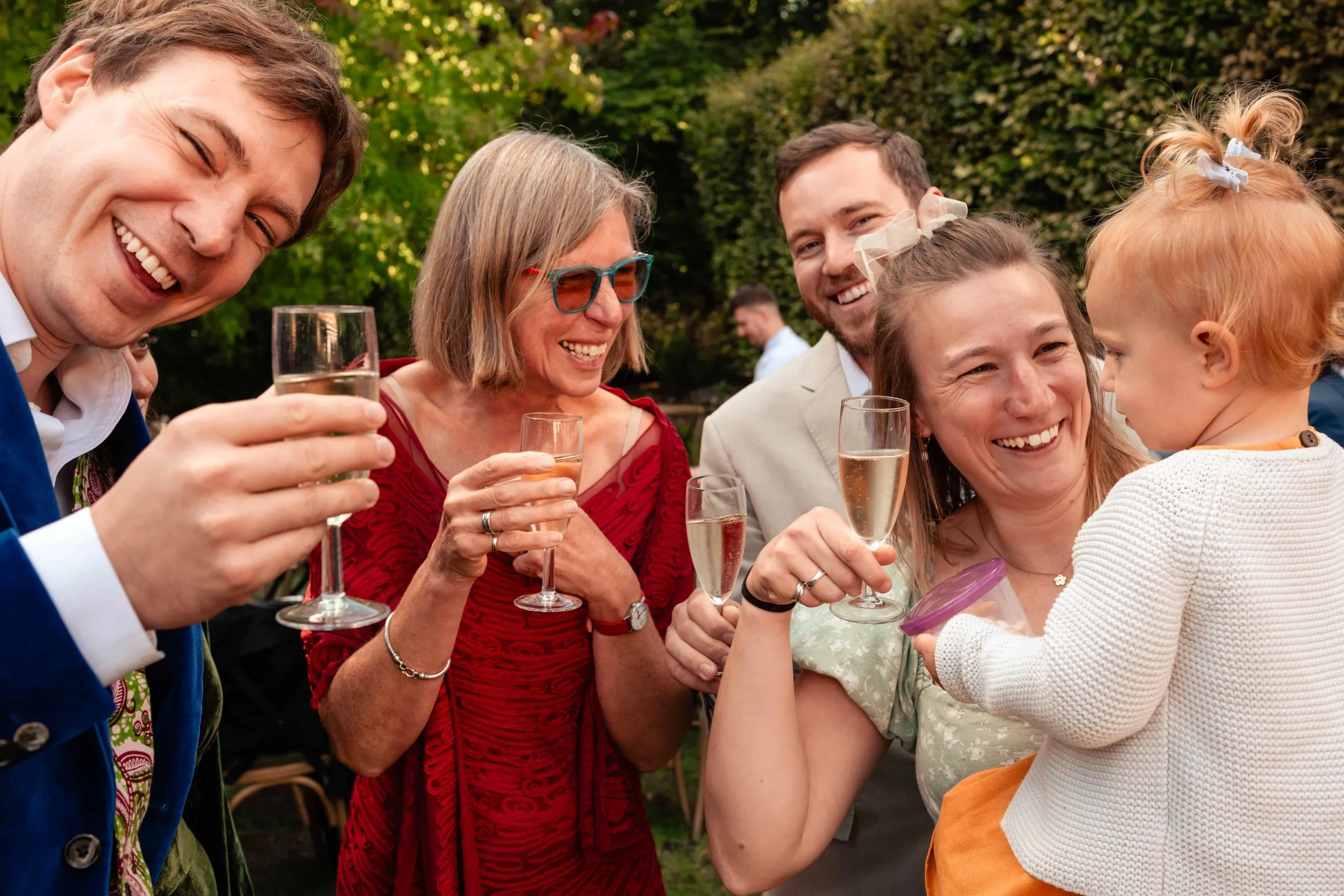 Group of five adults and one child enjoying a outdoor celebration, holding glasses of champagne and smiling, with green trees in the background.