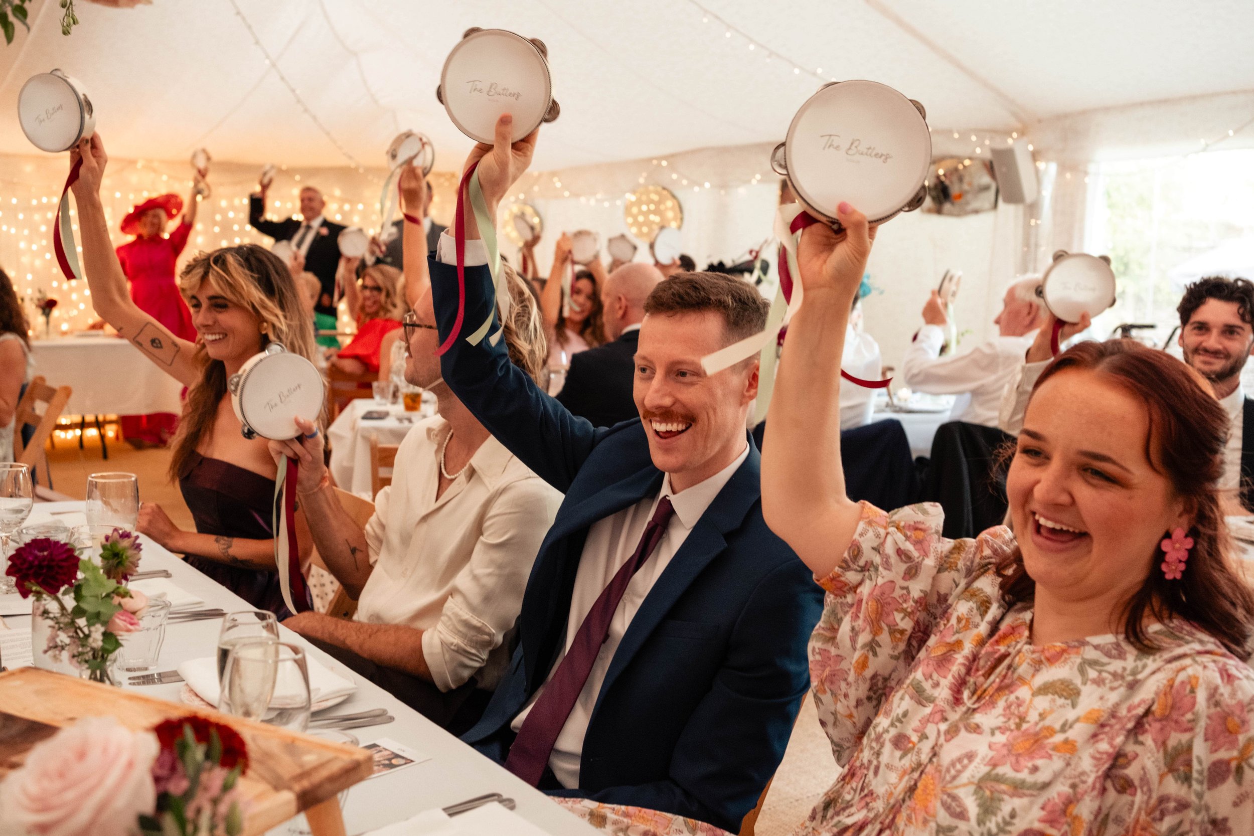 People at a celebration event, sitting at a banquet table, holding small musical drums with ribbons, smiling and raising them, decorated with flowers and string lights.