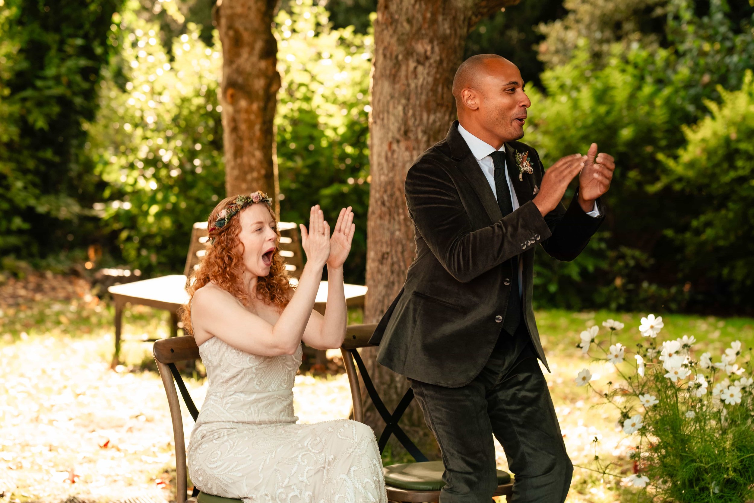 A woman in a white dress sitting on a chair outdoors with her hands raised and mouth open, appearing surprised or excited, while a man in a dark jacket and tie stands in front of her, clapping, during a daytime event in a wooded area with trees and g