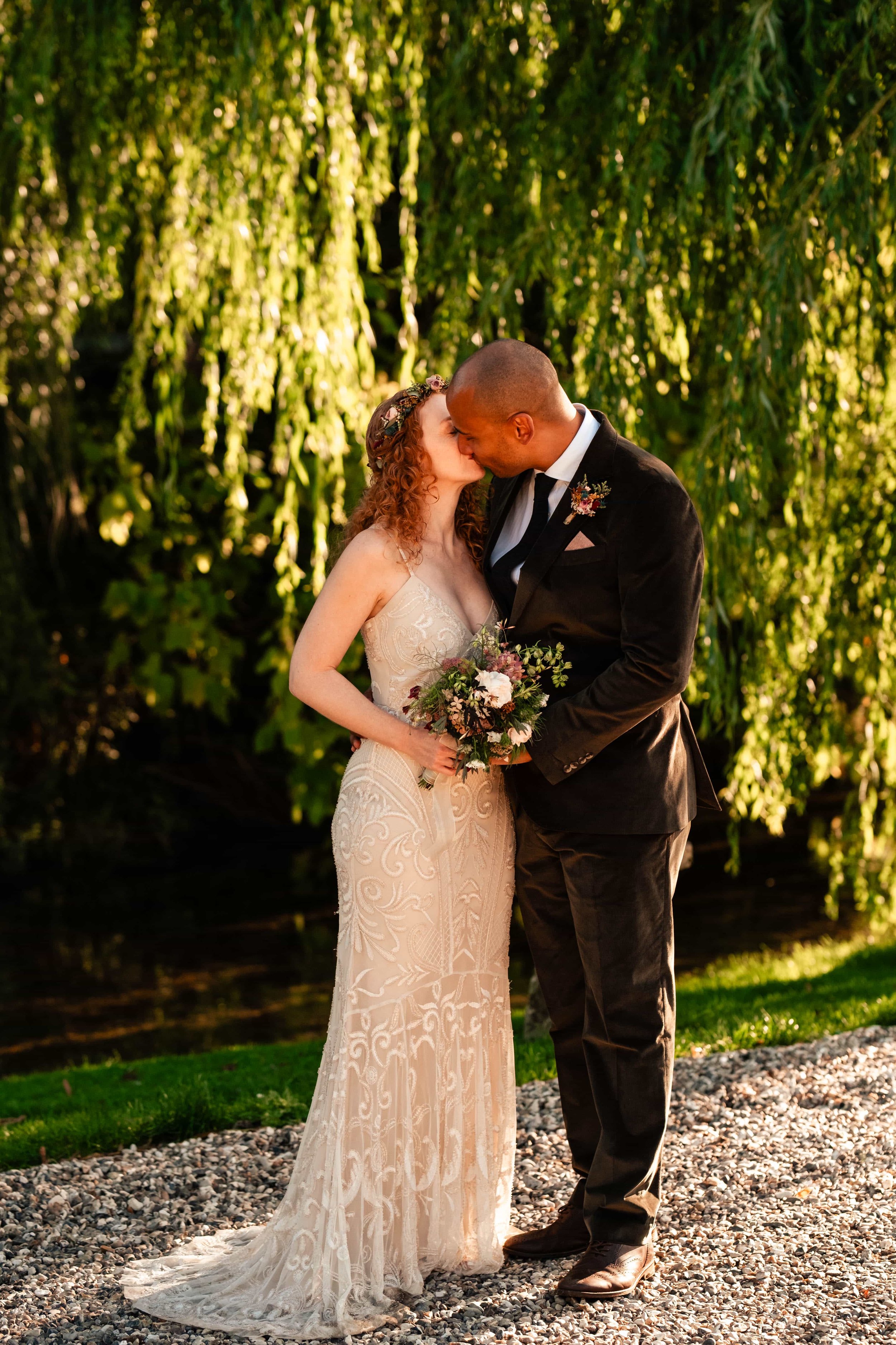 A newlywed couple sharing a kiss outdoors during sunset, with the bride holding a bouquet of flowers, surrounded by lush greenery.