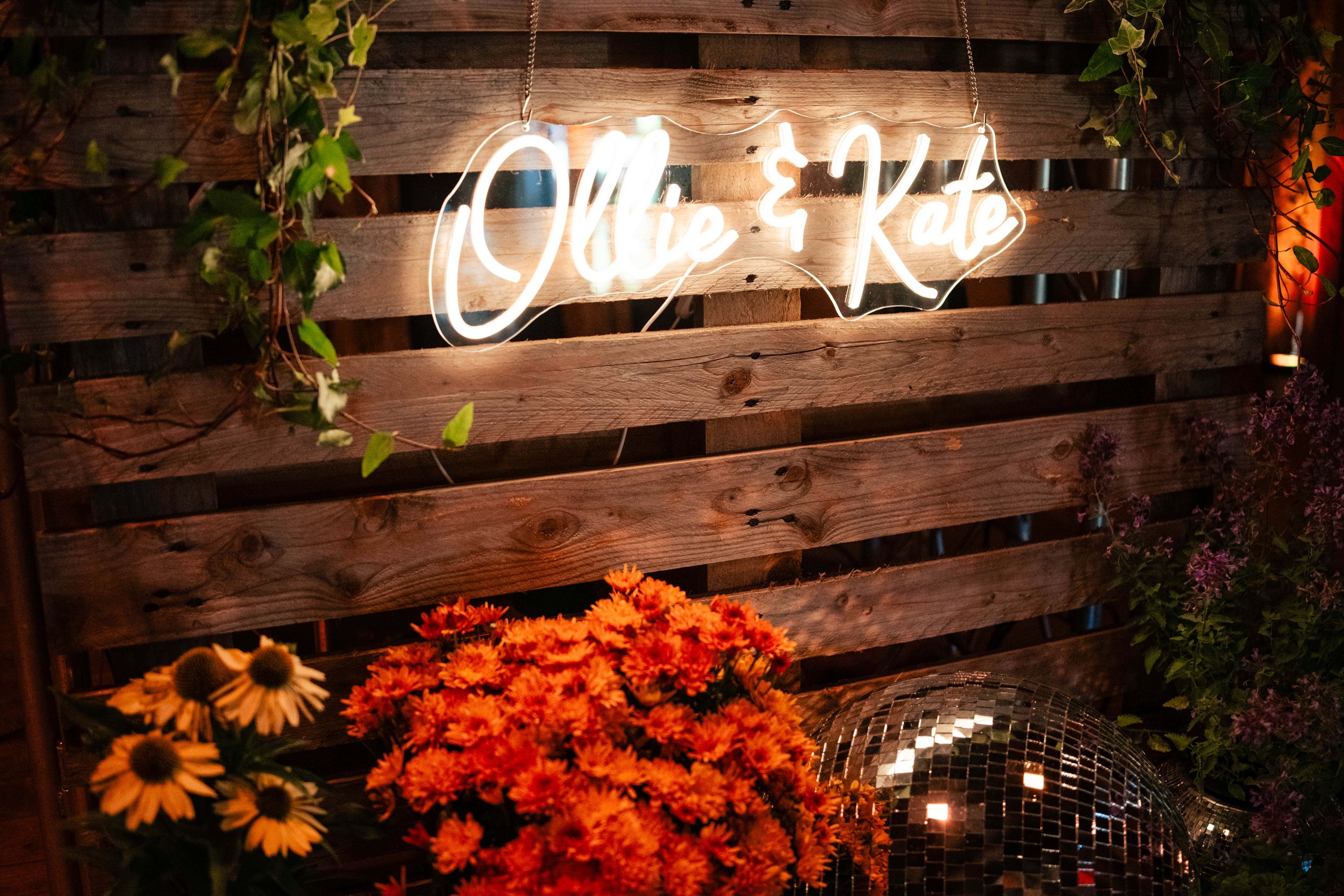 Neon sign reading 'Ollie & Kate' hanging above a wooden fence, surrounded by flowers and a disco ball.