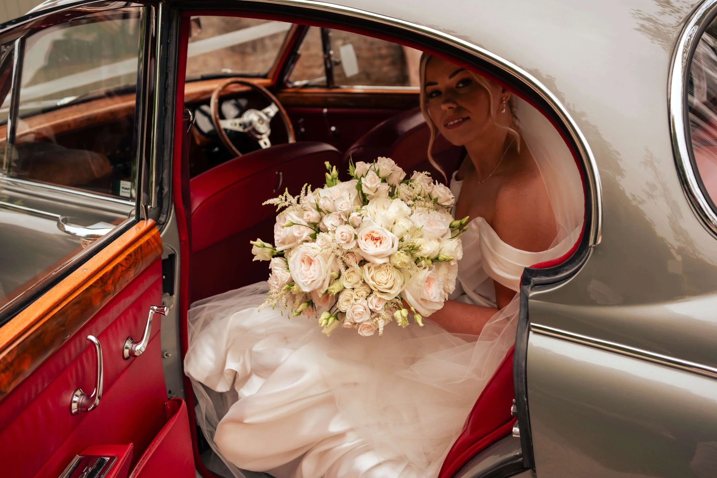 Bride sitting inside a vintage silver car, holding a large bouquet of white and blush roses, wearing a white wedding dress and veil.