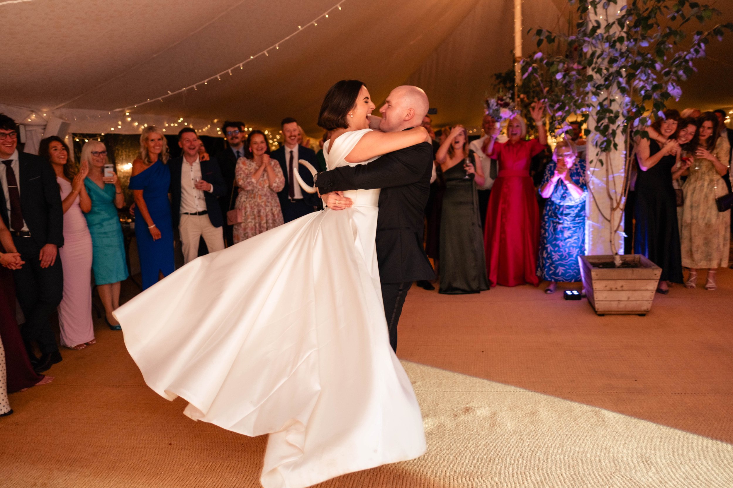 A bride and groom dance at their wedding reception, with guests watching and smiling in the background under warm lighting and fairy lights.