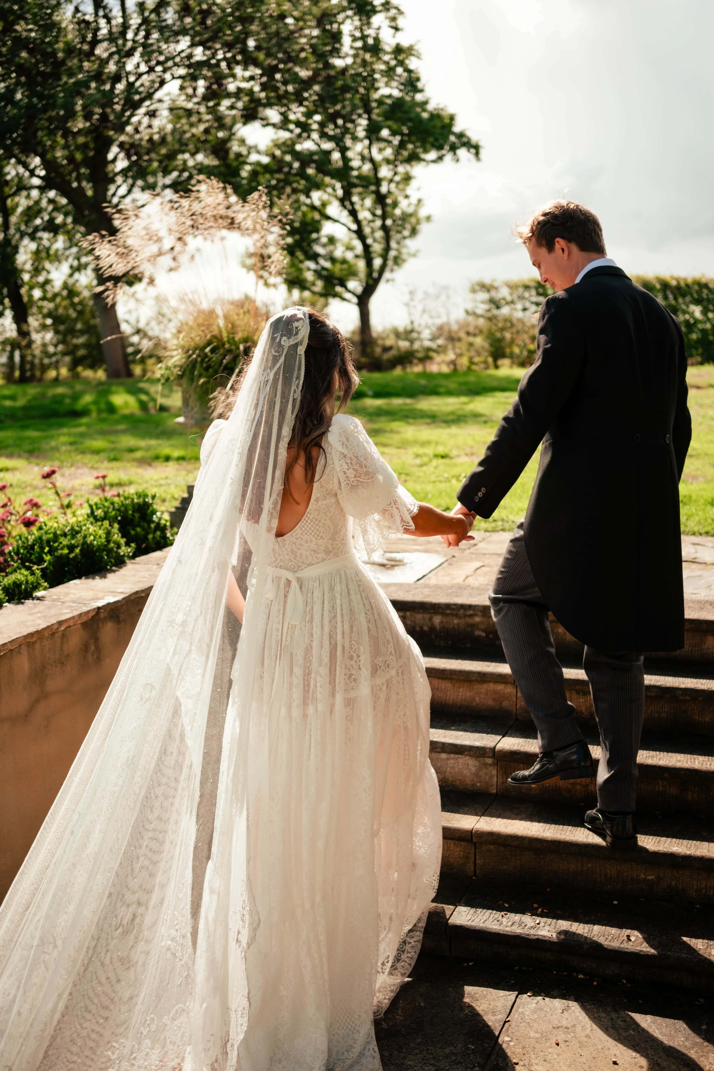 A bride and groom holding hands as they walk up outdoor steps during their wedding, with trees and greenery in the background.