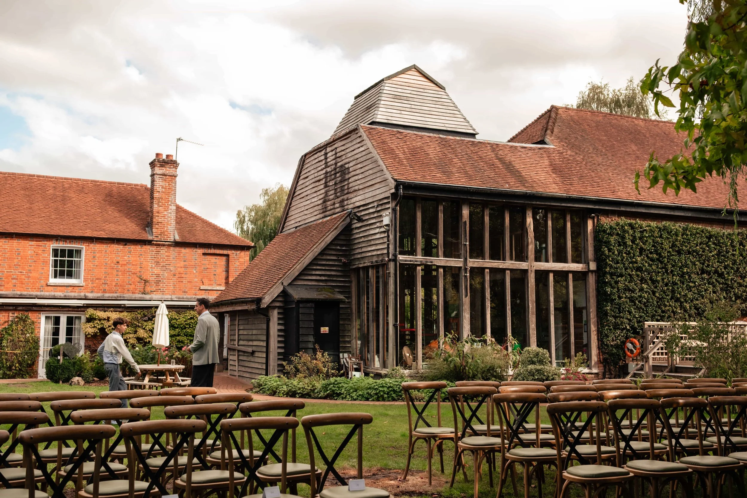 A rustic outdoor wedding setup with rows of chairs on a grassy lawn facing a wooden barn with a glass extension, two men talking, one young and one older, in front of the barn on a cloudy day.