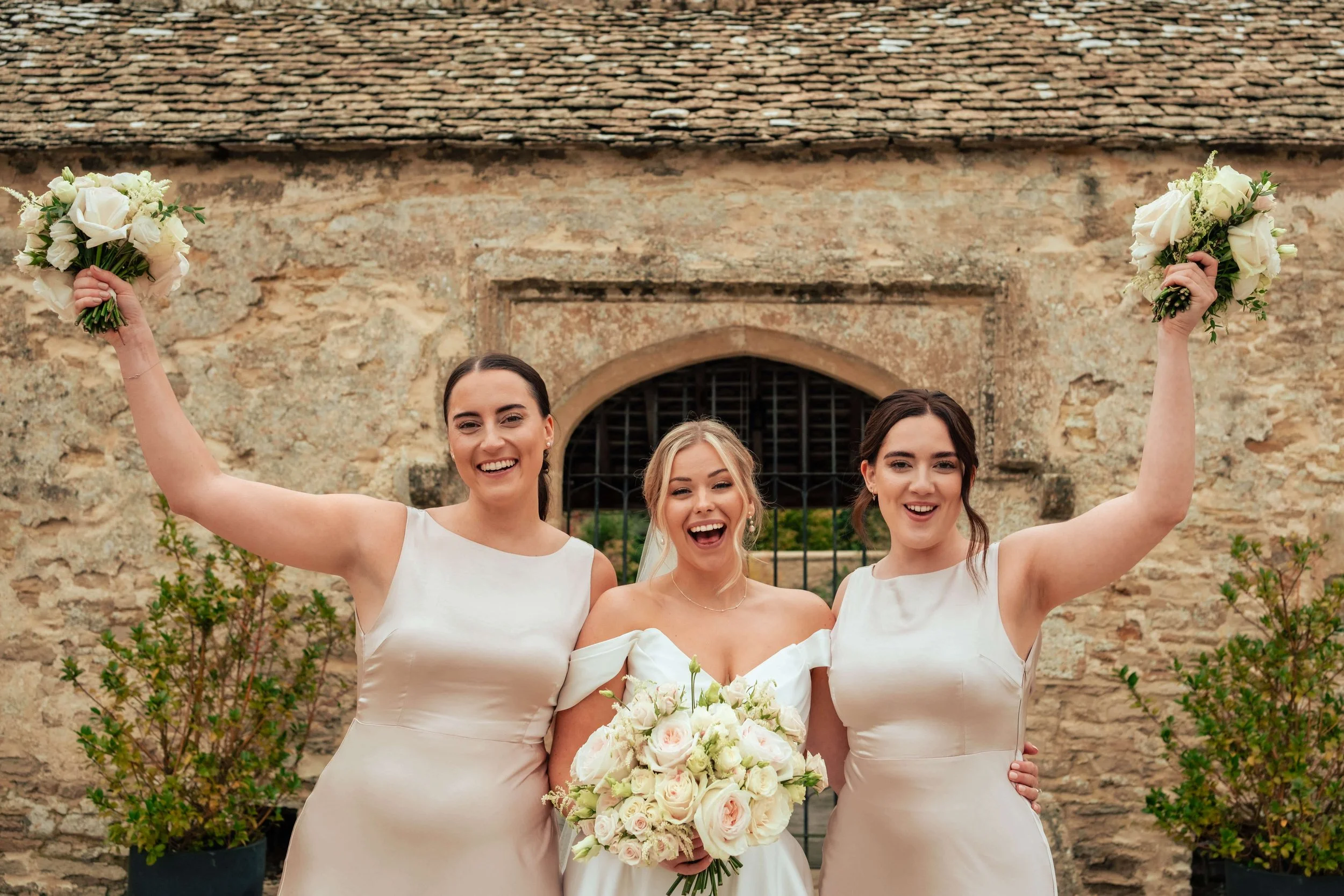 Three women dressed in white wedding attire smiling outdoors, with one in the middle holding a large bouquet of white and pink roses, flanked by two women holding smaller bouquets, standing in front of a rustic stone wall.