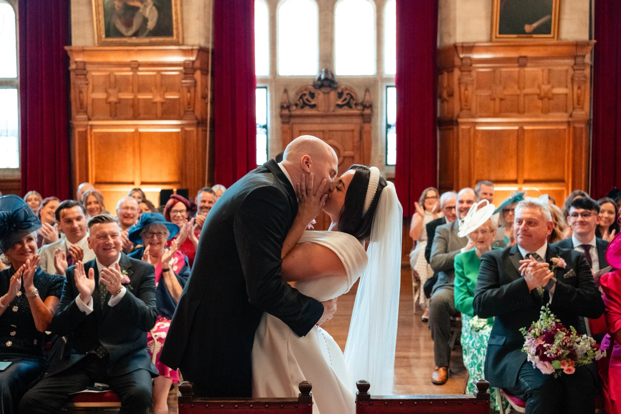 A couple's wedding kiss in a grand, wood-paneled room with red curtains, with guests clapping and smiling around them.