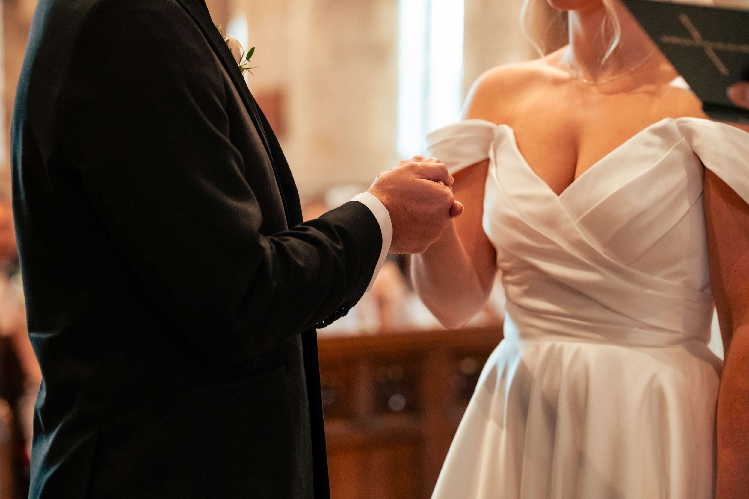 A couple getting married, holding hands during a wedding ceremony, with the bride wearing a white off-shoulder wedding dress and the groom in a black suit.