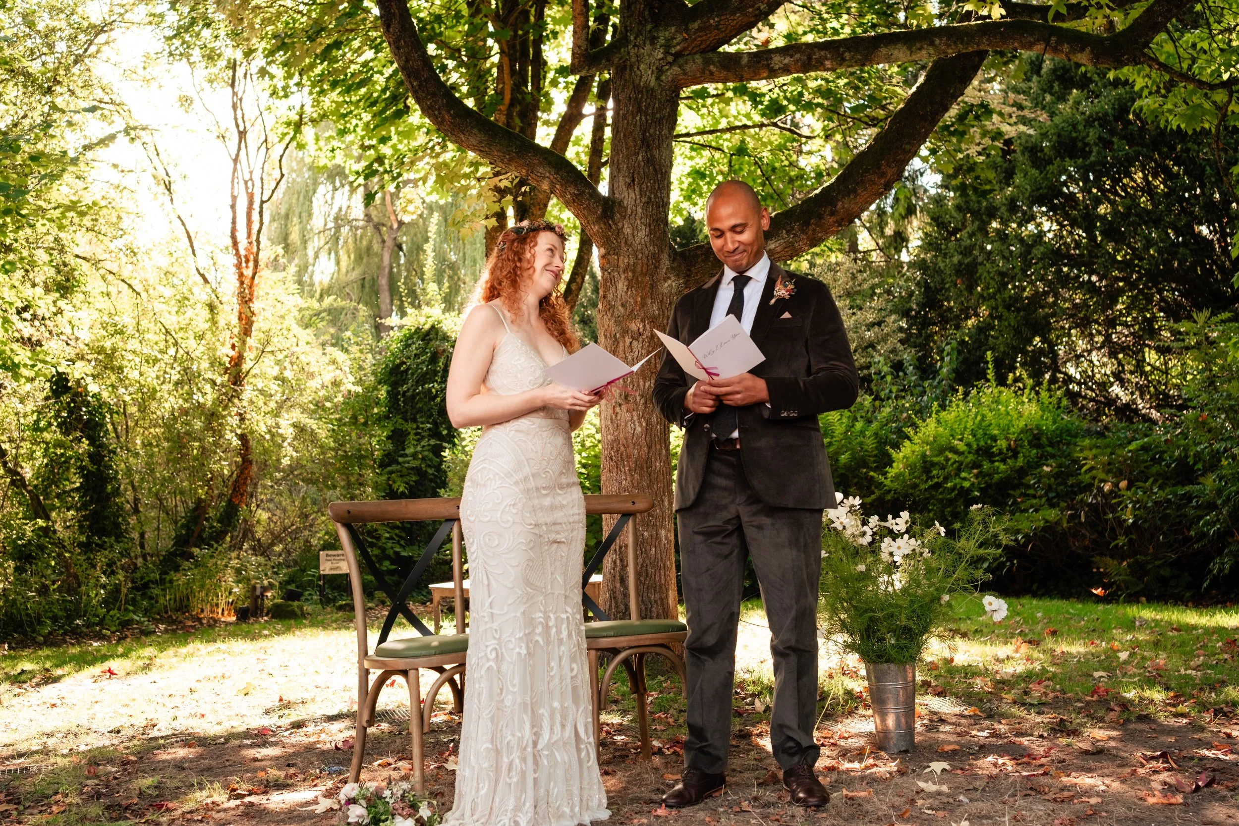 A couple exchanges vows outdoors during a wedding ceremony beneath a large tree with green foliage, with a wooden bench and white flower arrangement nearby.