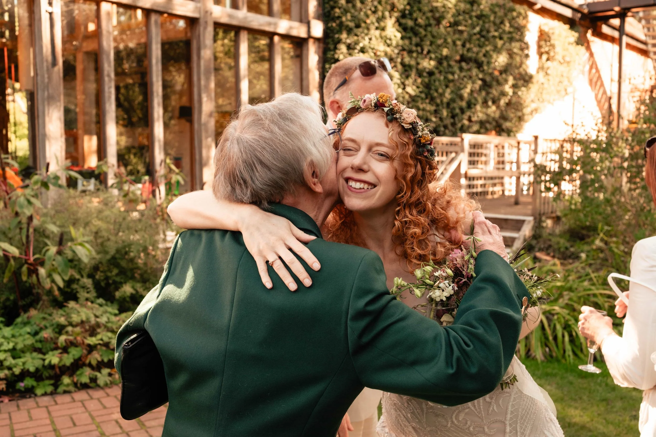 A young woman with curly red hair, wearing a floral crown and wedding dress, receiving a hug and kiss from an older man in a formal green suit at an outdoor wedding reception, with greenery and a wooden structure in the background.