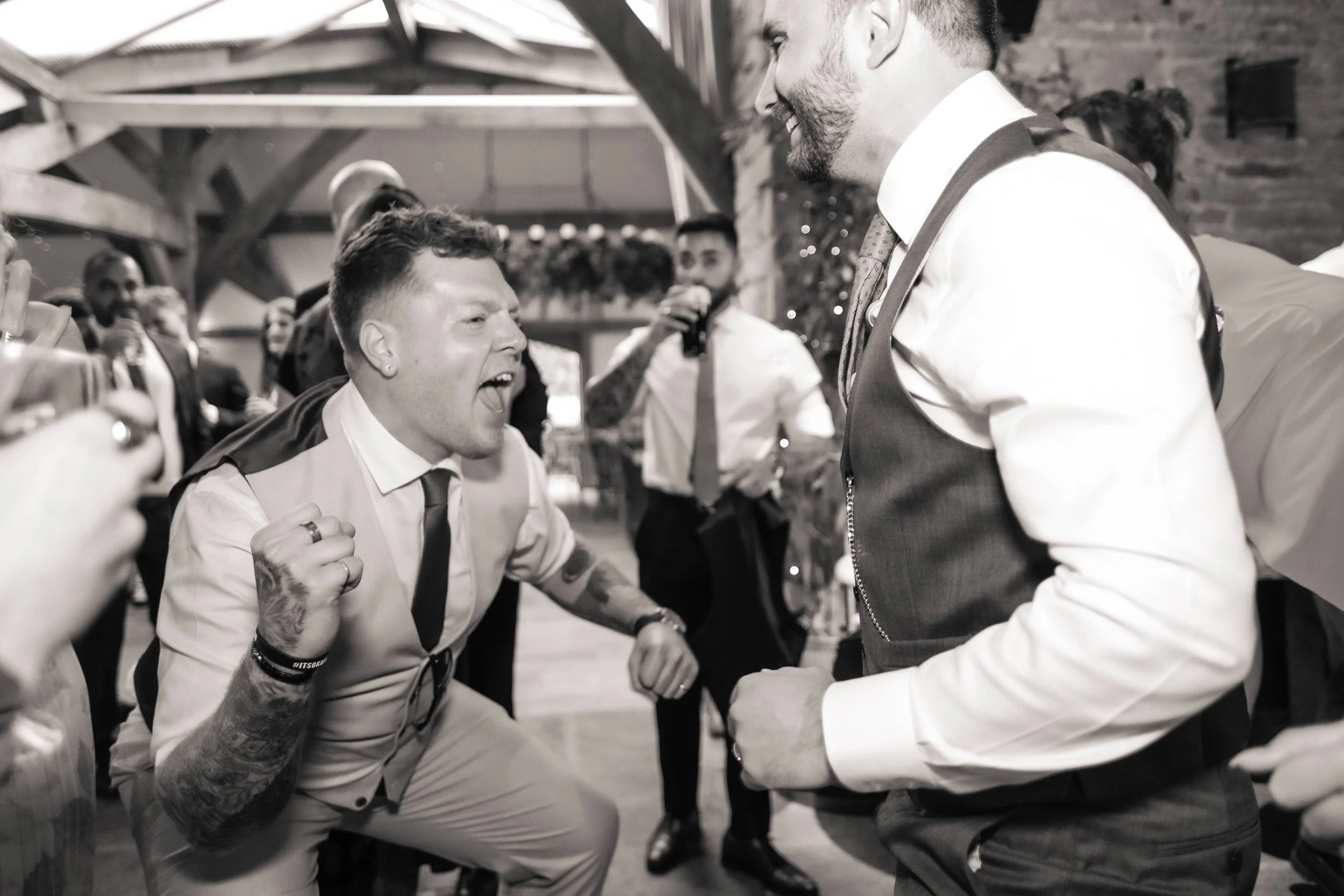 People dancing and celebrating at a lively indoor event, with one man passionately singing or shouting and another smiling and dancing passionately, surrounded by other guests in formal attire.