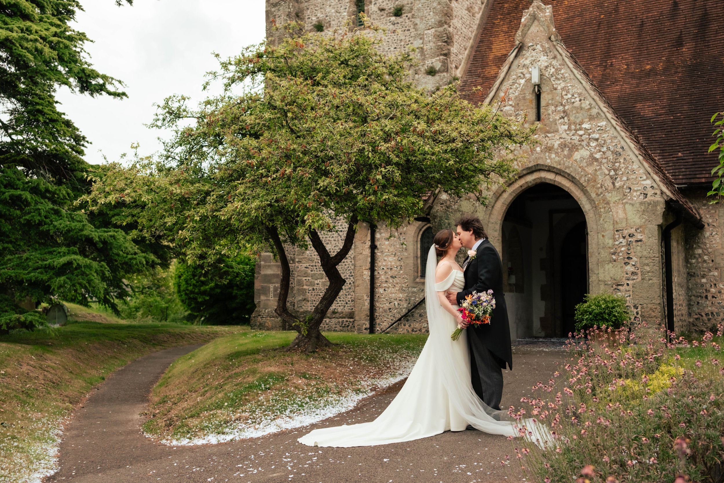 Bride and groom sharing a kiss outside the front entrance of the church, with confetti lining the pathway around them.