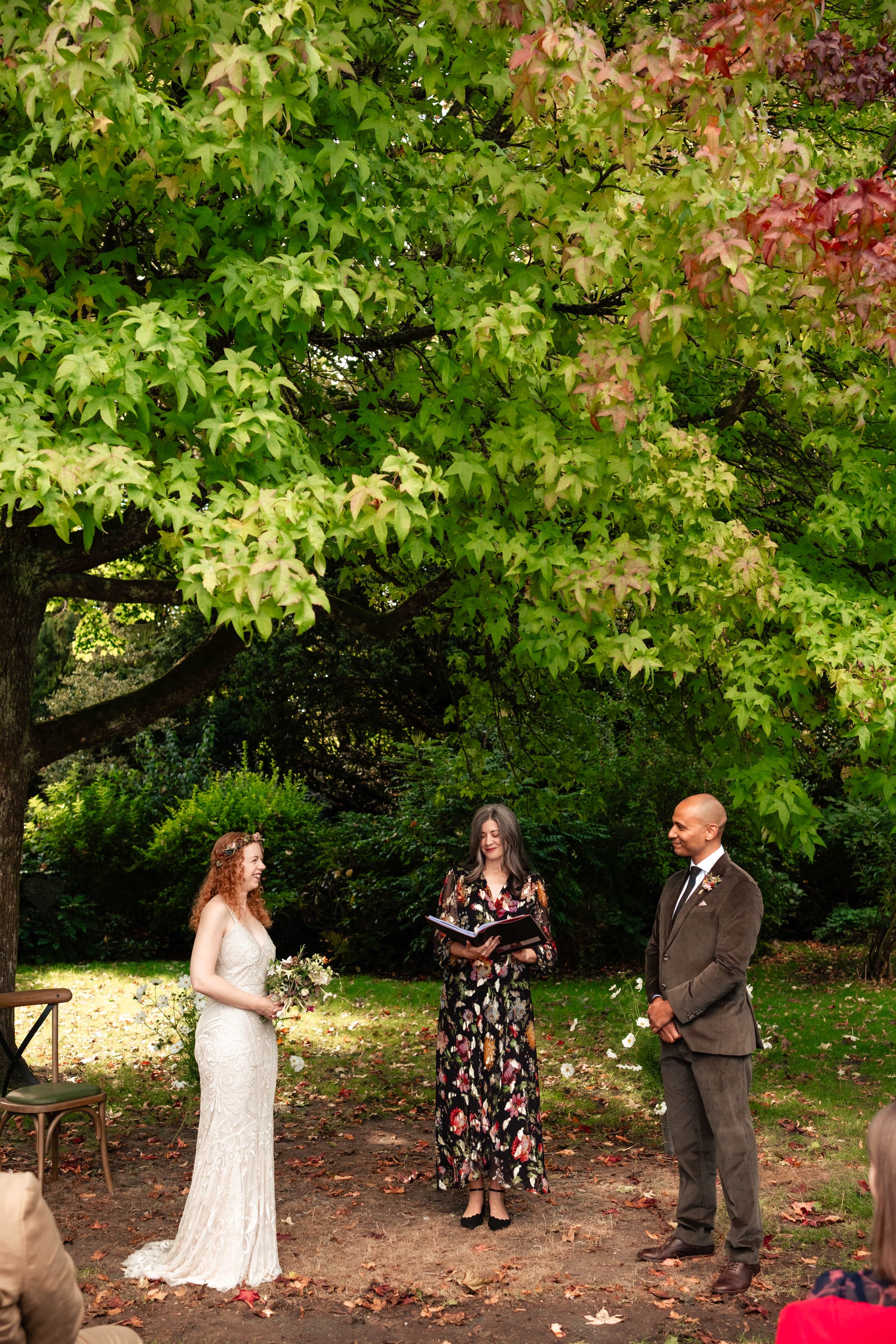 A wedding ceremony outdoors under a large leafy tree with three people: a bride with long curly red hair in a white lace wedding dress holding a bouquet, a groom with a bald head in a brown suit, and a woman in a black floral dress reading from a boo