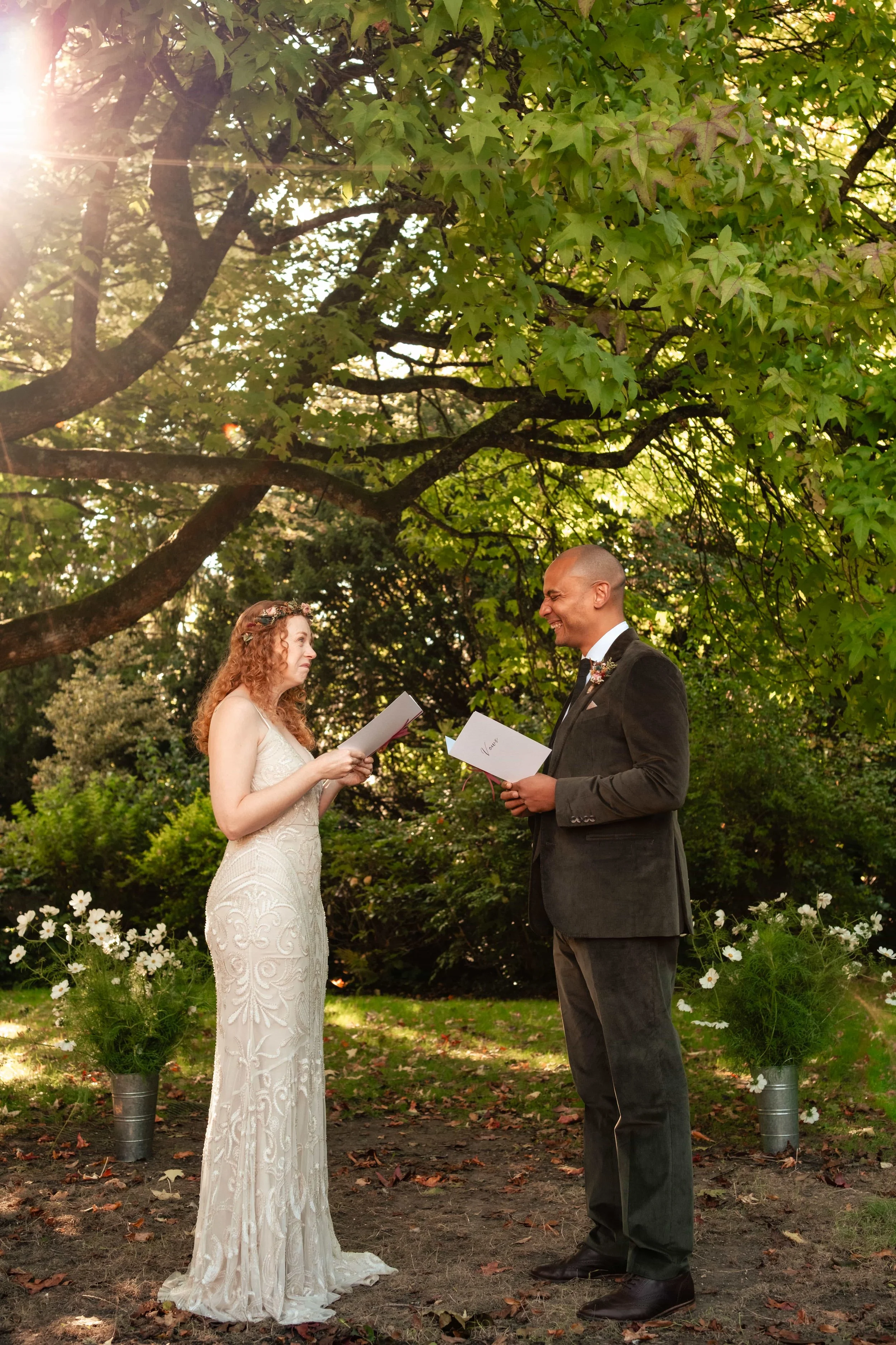A couple gets married outdoors under a large green tree, with sunlight filtering through the leaves. The bride has curly red hair and is wearing a white lace wedding dress, holding a paper, and the groom, a bald man in a dark suit with a boutonniere,