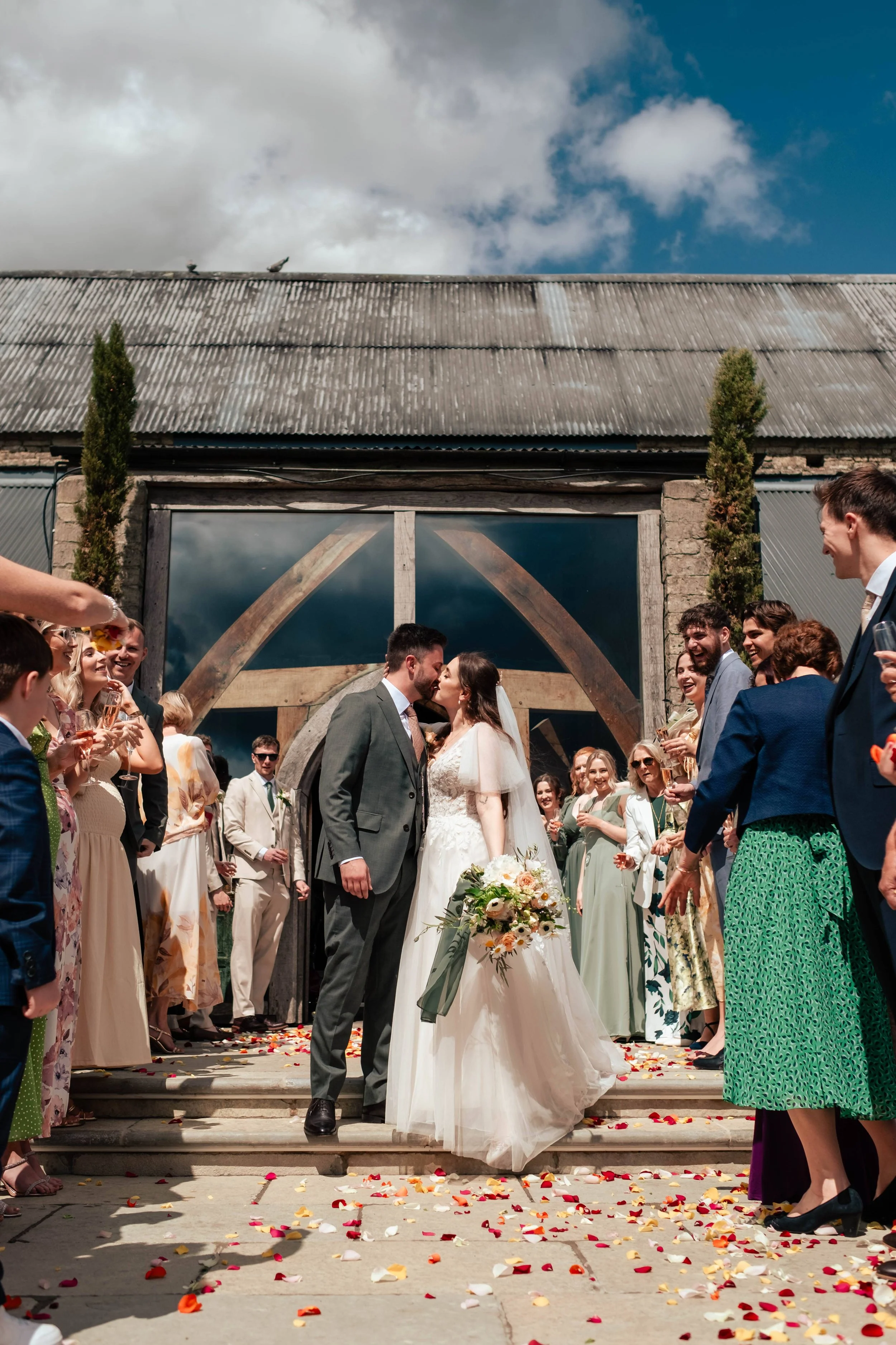 A wedding kiss between a bride and groom surrounded by friends and family outside a rustic barn, with rose petals scattered on the ground and a partly cloudy sky above.