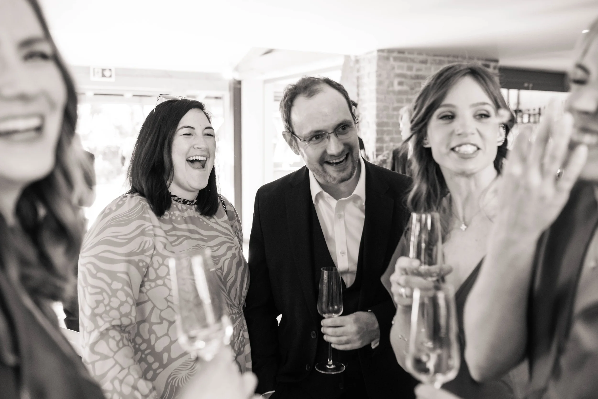 Group of people at a celebration, smiling and laughing, holding champagne glasses, in an indoor setting with exposed brick walls.